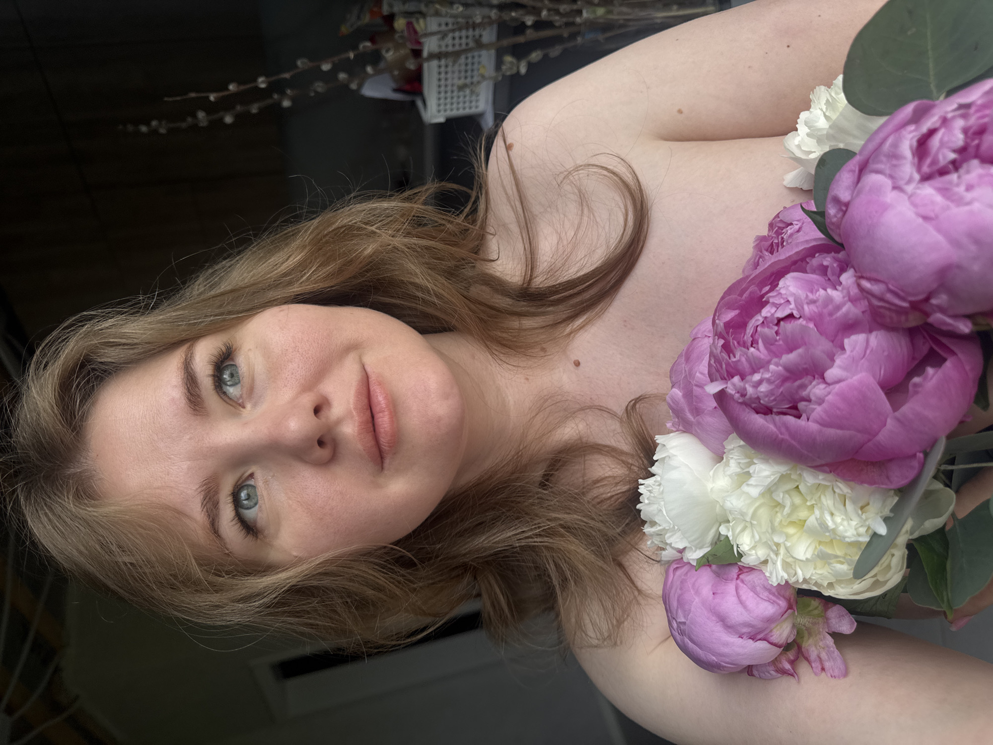 The image is a close-up portrait of a young woman, with the photograph rotated 90 degrees counter-clockwise. The woman appears to be lying down or reclining, looking towards the camera with a serene and softly smiling expression. She has light-colored, wavy hair (blonde or light brown) that frames her face and flows over her shoulders; a distinct strand on her bare shoulder forms a heart shape. Her fair skin features natural freckles and a few moles, and she has striking light blue eyes.
A vibrant bouquet of flowers rests on her chest and shoulder area, partially covering her upper body. The arrangement is dominated by large, lush pink peonies and smaller, ruffled white flowers, possibly carnations, accented with green foliage, likely eucalyptus leaves. Both the woman's face and the flowers are in sharp focus.
The setting appears to be indoors, with a dark, blurred, and out-of-focus background creating a shallow depth of field. Subtle details in the background include fuzzy pussy willow branches, suggesting a springtime atmosphere. The lighting is soft and diffused, contributing to a gentle and intimate mood. There is no visible text in the image. The scene captures a moment of calm and natural beauty, located in an indoor setting in Ферма, Russia.