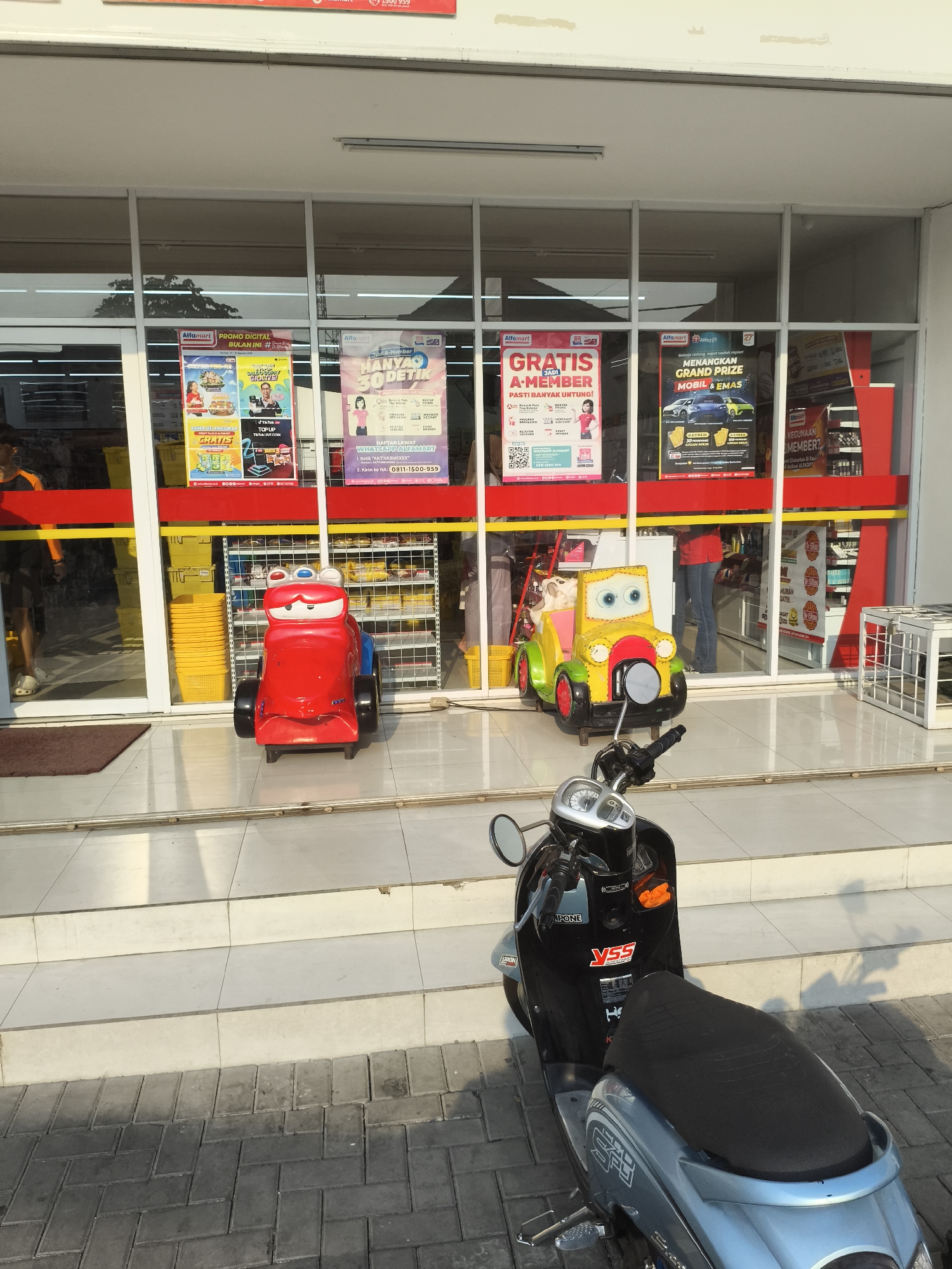 The image depicts the exterior of an Alfamart convenience store in Depok, Indonesia. In front of the store's entrance, there are two children's ride-on toys: a red one resembling the character Lightning McQueen from the movie "Cars," and a yellow and green toy car. To the right, a black and silver scooter is parked on the tiled sidewalk. The store has large glass windows displaying various promotional posters, including ones for "Promo Digital," "Gratis A-Member," and "Menangkan Grand Prize Mobil & Emas." Inside the store, shelves stocked with products are visible. The overall scene suggests a typical commercial street view in Depok.