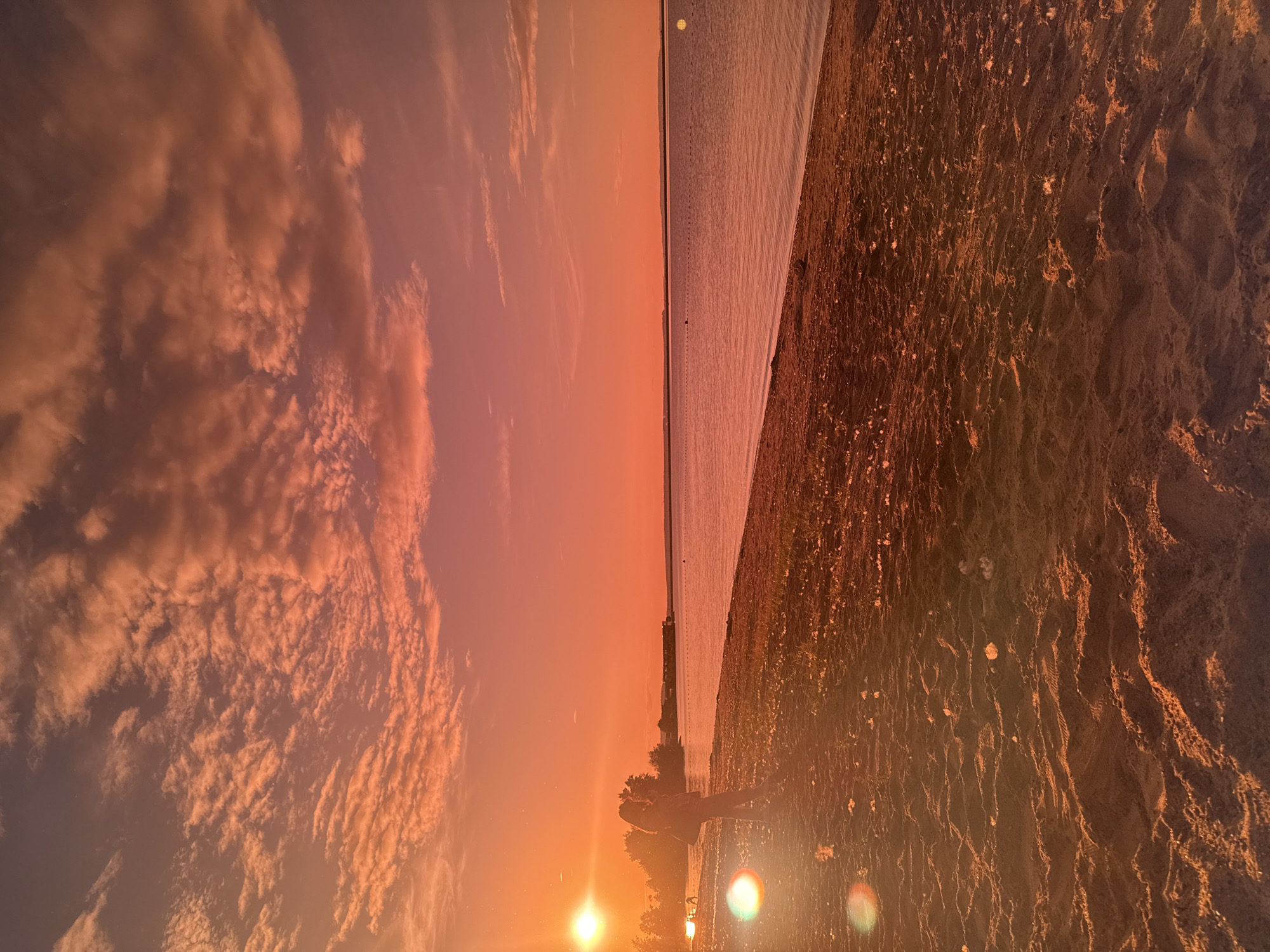 The image captures a sunset scene over a large body of water in Chaikovsky, Russia. The sky dominates the upper two-thirds of the frame, displaying a vivid gradient of orange, red, and pink hues, with numerous scattered clouds illuminated from below. The sun, a brilliant orb, is positioned low on the far left horizon, generating a prominent lens flare in the lower-left foreground.
A sandy beach forms the foreground, stretching from the bottom center towards the middle right. The sand exhibits textural ripples, depressions, and scattered footprints. Some light-colored detritus, possibly small shells or dried vegetation, is visible on the sand.
A solitary individual, appearing female, stands in profile on the beach in the mid-left, facing towards the right. They are largely silhouetted against the bright setting sun but appear to be wearing long pants and a top, with long hair visible. Their left arm is bent, holding an indistinct object.
A vast, calm body of water occupies the middle ground and background, reflecting the warm colors of the sky in muted tones. A horizontal line of small, dark buoys stretches across the water in the mid-distance. A low-lying landmass or distant shoreline is faintly visible on the far horizon across the water, under the brightly colored sky.