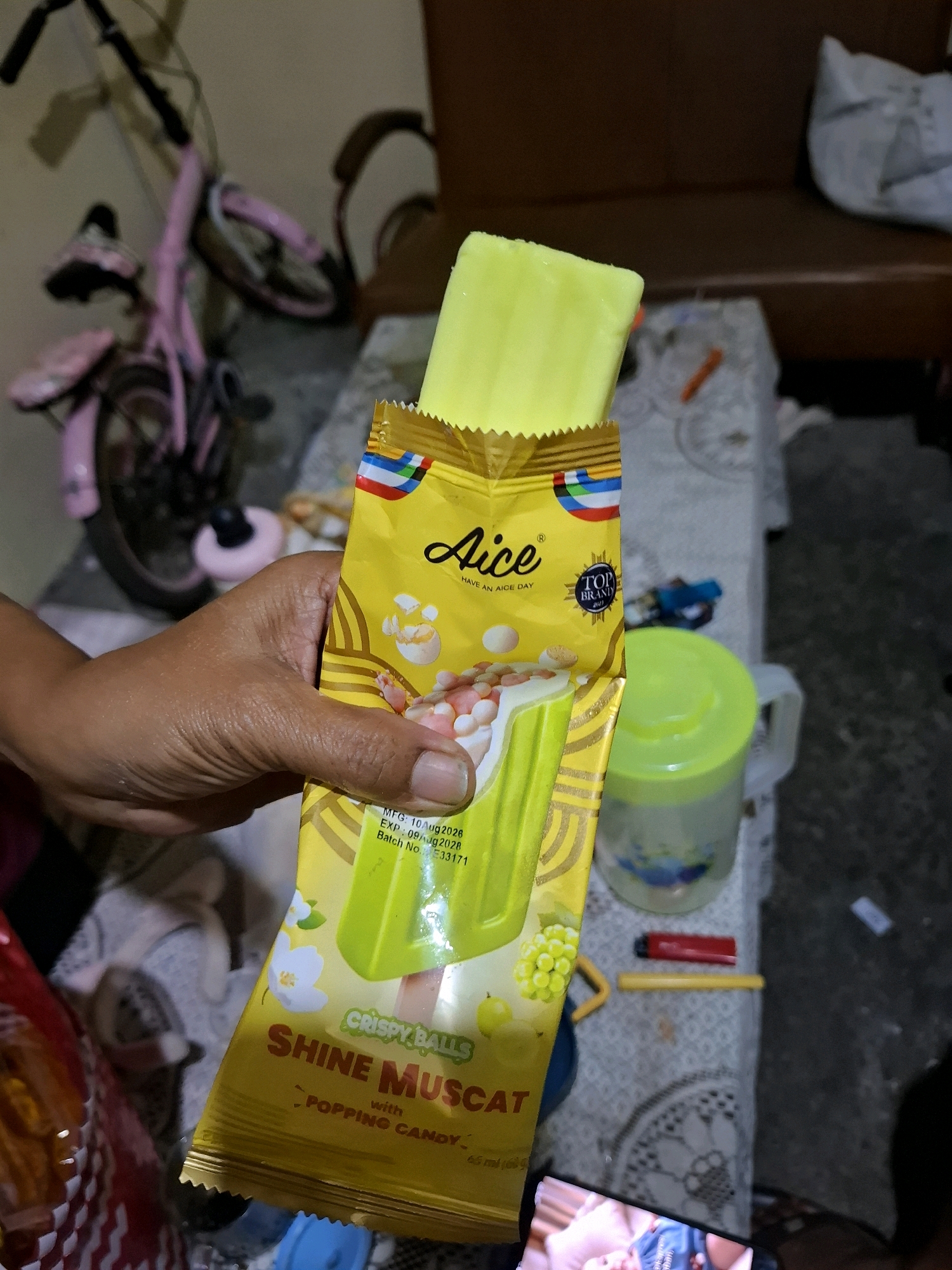 A person's hand holds a partially unwrapped Aice Shine Muscat ice cream bar. The ice cream is light green and appears to be a popsicle. The wrapper is primarily yellow with red, blue, and white accents. Text on the wrapper indicates it is "CRISPY BALLS SHINE MUSCAT with POPPING CANDY". The background is blurred but suggests an indoor setting, possibly a home, with a pink bicycle visible to the left, a wooden bench or sofa in the background, and a patterned tablecloth on a table. A green plastic pitcher and a red lighter are also on the table. The lighting is dim.