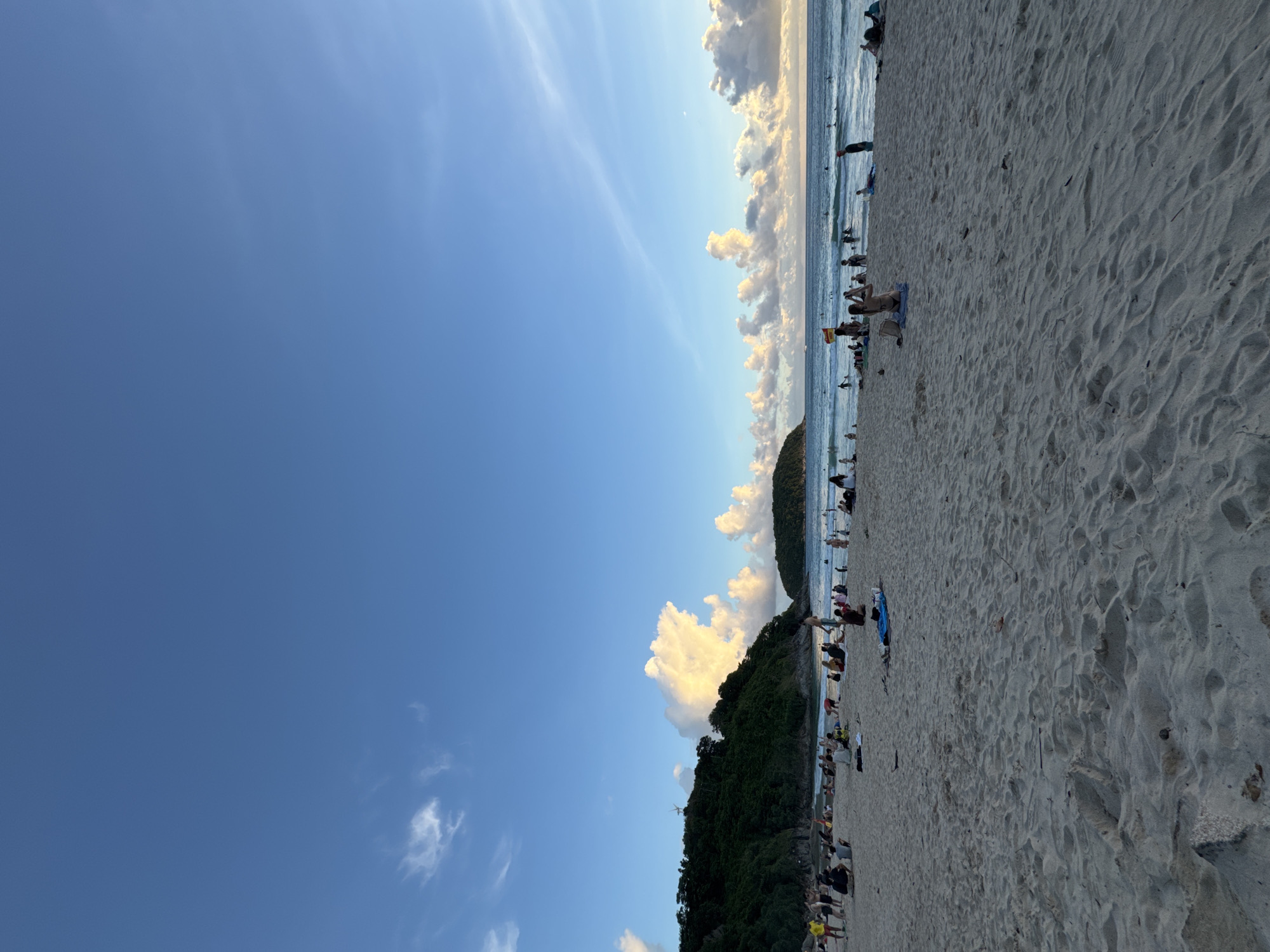 The image depicts a crowded beach scene in Rawai, Thailand, likely during sunset given the lighting. The foreground is dominated by sand, with footprints and some scattered debris. In the midground, numerous people are seated or lying on the sand, some on towels or mats, and others are walking or standing. The middle ground also shows the edge of the ocean with gentle waves, and several individuals are in the water, some with surfboards.
Further back, a lush green, tree-covered hill forms the left side of the scene, with a small island visible in the distance to the right. The sky is a vibrant blue with scattered, puffy white clouds, some illuminated by the setting sun. The overall impression is a lively, recreational atmosphere on a tropical beach.