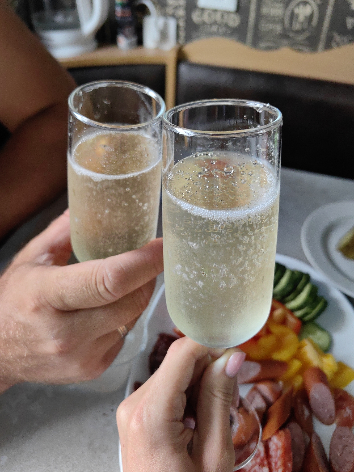 Two glasses of sparkling wine are held by two people. The glasses are tall and narrow, filled with a pale yellow bubbly liquid. A plate of sliced sausages, peppers, and cucumbers is visible in the background. The setting appears to be a casual outdoor or indoor space, possibly a cafe or restaurant. The image does not provide any information about the city of Fanipal, Belarus.