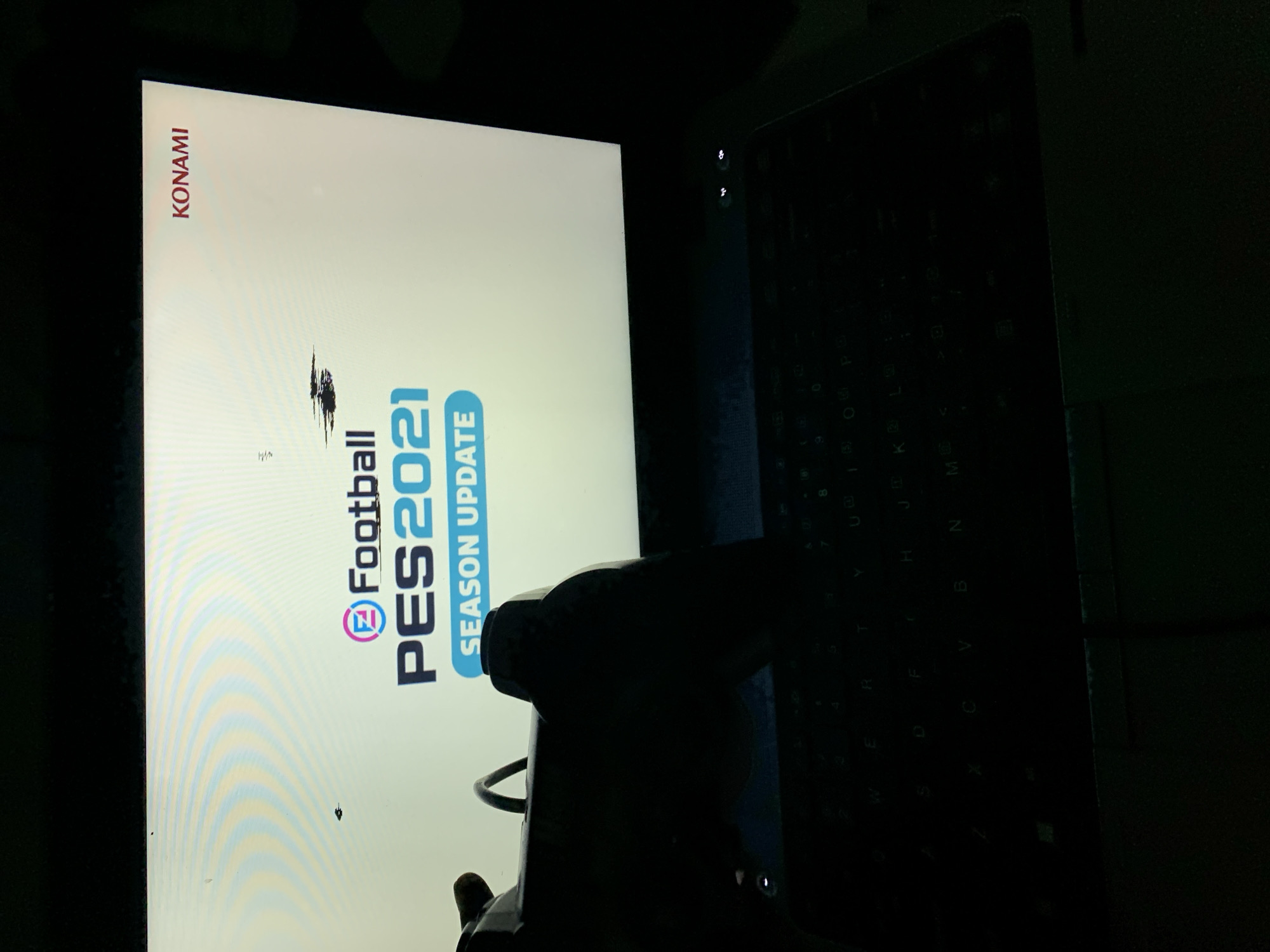 Here's a description of the image:
**Scene:** A laptop screen displays the title screen of the video game "eFootball PES 2021 SEASON UPDATE". The word "KONAMI" is vertically aligned at the top left of the screen. A PlayStation gamepad sits on top of the laptop keyboard.
**Environment:** The surrounding environment is dark.