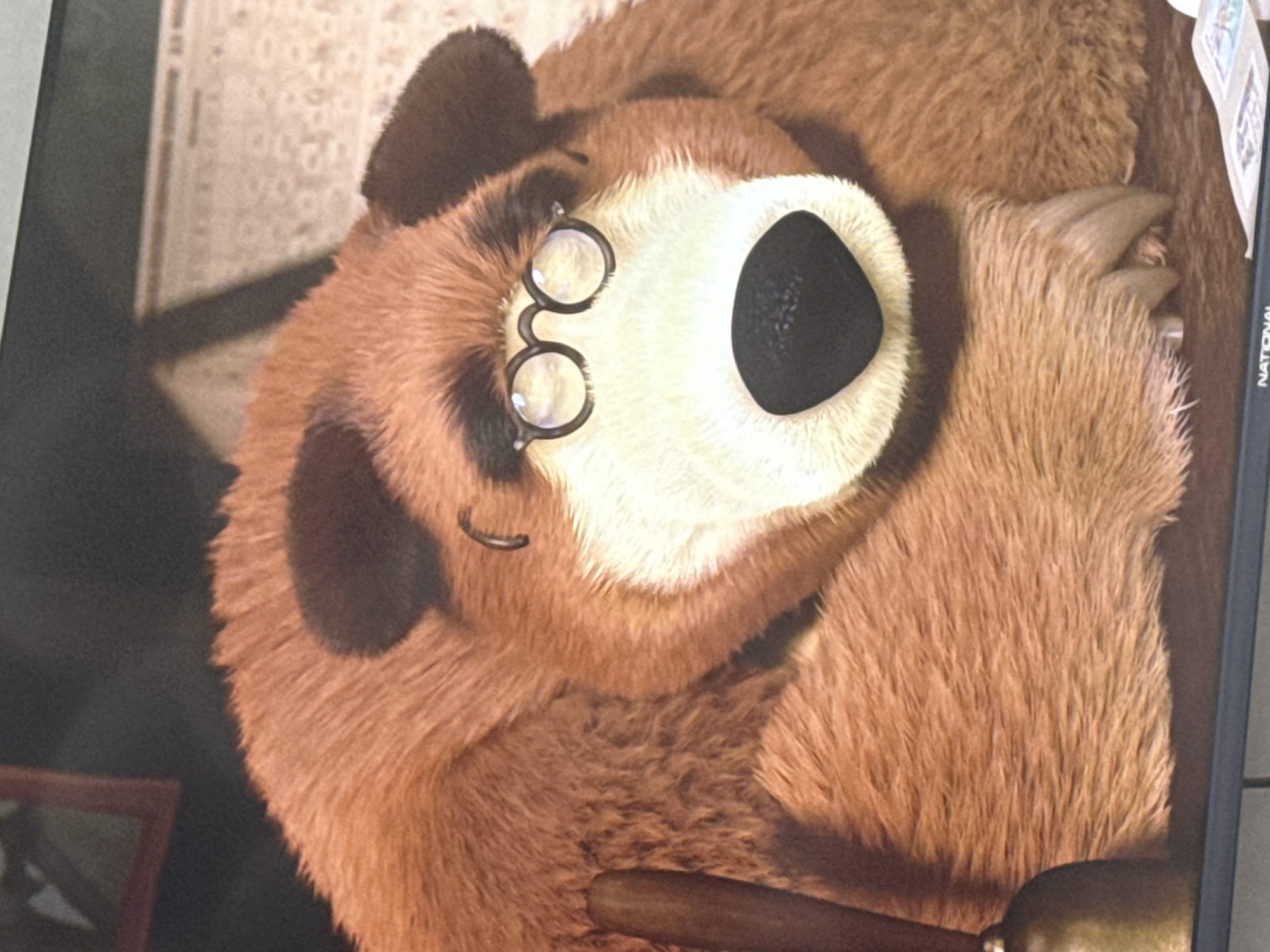 This media file shows a close-up of a stuffed toy bear. The bear is brown with darker brown markings around its eyes and ears. It is wearing small, round, wire-rimmed glasses. The bear's nose is a large, black, oval shape. Its fur appears soft and fluffy. One of the bear's paws is visible, with blunt claws. The bear is lying down, with its head tilted upwards. In the background, there are hints of a room, including what appears to be a patterned wallpaper and possibly a wooden frame or furniture. Part of a dark object, possibly a gavel, is visible in the foreground, resting on the bear. A narrow, dark vertical element with some text is visible on the right side of the frame. The lighting suggests an indoor setting. The image does not provide information about the time of day, weather, or specific emotions, other than the gentle and perhaps slightly whimsical appearance of the toy. No specific location cues for Yousovo, Russia are evident within the image itself. The text on the right side of the image includes the word "NATIONAL" in capital letters.