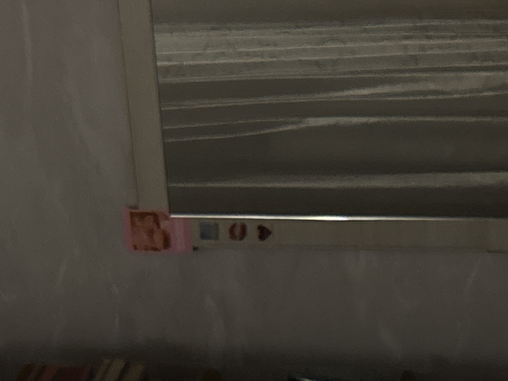 The image presents a dimly lit, blurry indoor scene, focusing on a large, rectangular reflective surface, likely a mirror or a window with horizontal blinds/shutters. The surface occupies the right and central upper portion of the frame, showing faint horizontal lines that could be reflections of blinds or the blinds themselves.
To the left, a light-colored wall, possibly beige or grey, with a subtle texture, is visible. At the bottom left edge of the reflective surface, a small, pink, rectangular object is attached, possibly a sticker or a note. It displays an indistinct orange or brown image, possibly a stylized face or figure.
Adjacent to this pink object, on a metallic or white horizontal frame, are three small, colored symbols. From left to right, these are a light blue or grey square, a reddish-brown inverted 'U' or incomplete circle, and a dark red or brown triangle pointing right. These symbols resemble controls or indicators for an electronic device.
Below the reflective surface, the bottom edge of the image is very dark and out of focus, hinting at objects such as books or items on a shelf. No people are visible, and no specific activity is taking place. The overall dimness and blur suggest the photograph was taken in low light conditions, possibly evening or night, or in a poorly illuminated room. No legible text is discernible.