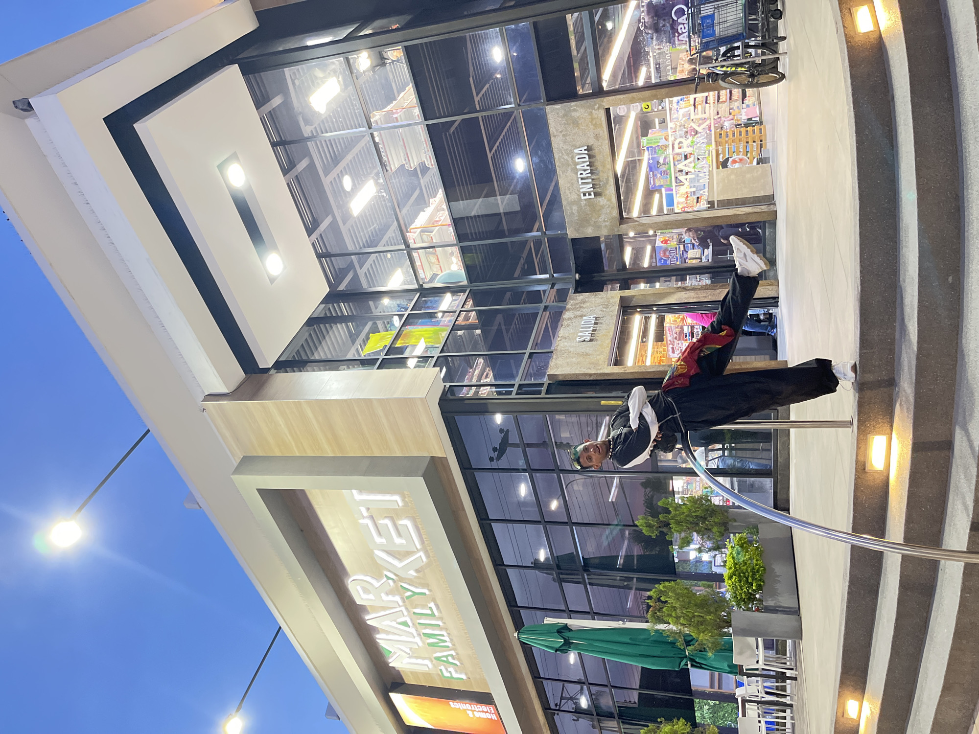 A rotated, low-angle exterior shot captures a person posing on an exterior railing outside a building. The building's facade features the words "FAMILY MARKET" stacked vertically on a light-colored sign with a contrasting border. The architecture is modern, with extensive glass windows reflecting interior views and the sky above. The building appears to be a grocery store or supermarket, with the word "ENTRADA" visible above an entrance. Various items, including shopping carts, are visible within the store. The sky is dusky, suggesting either dawn or dusk. The person is wearing dark clothing and white shoes, leaning backward on the railing. There are some small trees in pots and outdoor seating in front of the building. The location is plausibly the Family Market grocery store in Pampatar, Venezuela, based on the name and architectural style.