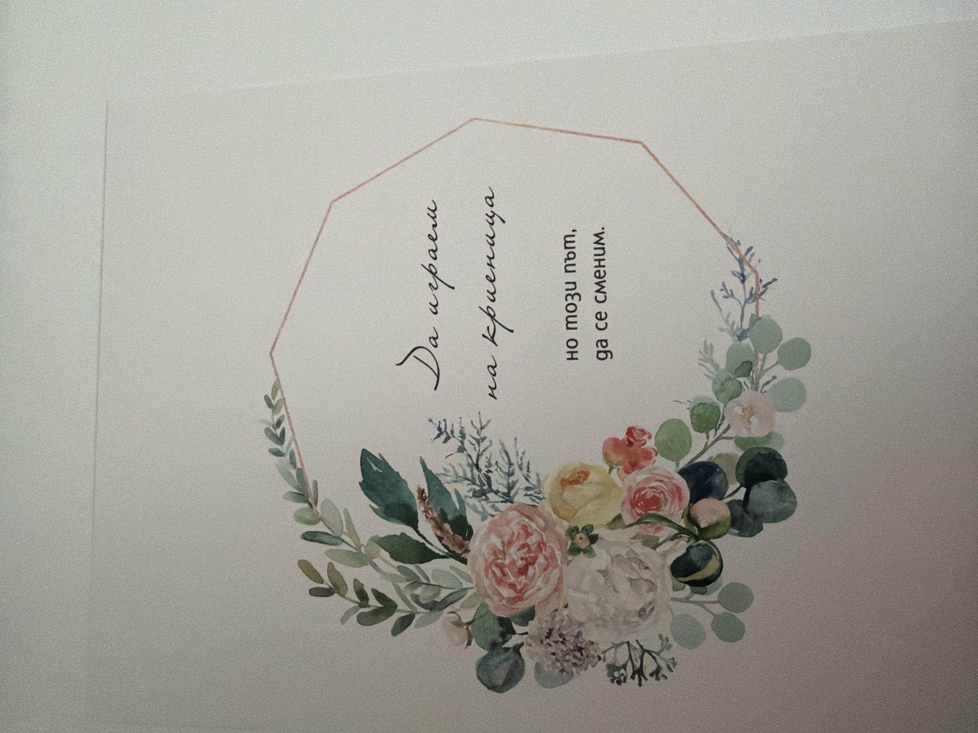 This is a close-up, eye-level, indoor photograph of a piece of paper with a floral watercolor design and text. The paper appears to be a card or a print. A delicate border of watercolor flowers and greenery, predominantly in shades of pink, white, yellow, and green, frames the left and top edges of the paper. A polygonal shape, outlined in a light peach color, is present in the center right of the image. Within this shape, there are three lines of handwritten text in Bulgarian. The text appears to be a quote or a message. The background is a plain, light-colored surface, likely a wall or a table, with subtle texture and slight shadowing indicating indoor lighting. There are no people or animals visible in the image. The photograph is taken with a slight angle, and the lighting casts subtle shadows. There is no information in the image that would indicate a specific location in the city of Pazardzhik, Bulgaria.