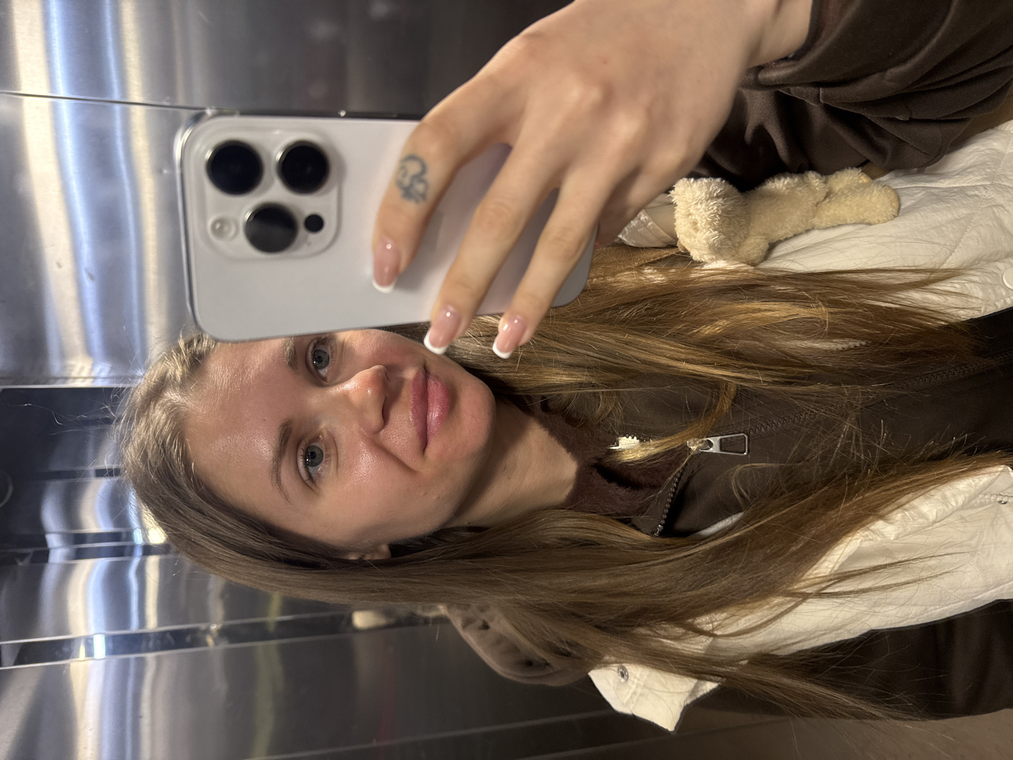 A woman is taking a selfie in a mirrored elevator. She is holding a silver iPhone in her right hand. Her fingernails are manicured with a white tip. There is a small tattoo on her right ring finger. She has long, light brown hair. She is wearing a brown zippered jacket and a white jacket underneath. Behind her, partially visible, is a small, tan, stuffed animal. The reflective surface of the elevator shows blurred details of the surroundings. There is no definitive information in the image to determine whether the location is in Batumi, Georgia.