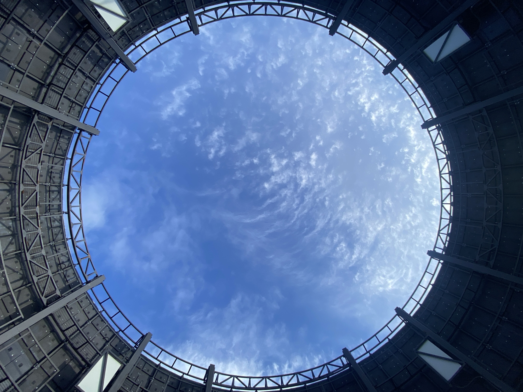 Looking upwards from within a circular, enclosed structure in Шебунино, Russia, a bright blue sky dotted with white cirrus clouds fills the opening. The structure appears to be made of metal and features a grid-like pattern, with some rectangular openings containing white or light-colored panels. The circular opening at the top is framed by the metal framework of the structure.