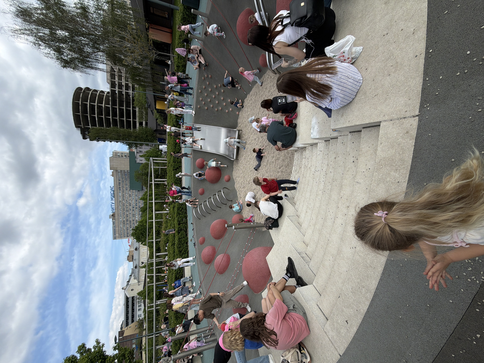 A playground scene, possibly in Moscow, Russia, shows numerous people, mostly children with a few adults, participating in activities. The setting includes a slide, red sphere-shaped structures, and climbing equipment. Some individuals are sitting on stairs, while others are playing. A tall, skeletal building stands in the background. The sky is cloudy with patches of blue. A building labeled "НИТУ МИСИС" is also visible. The image is rotated.