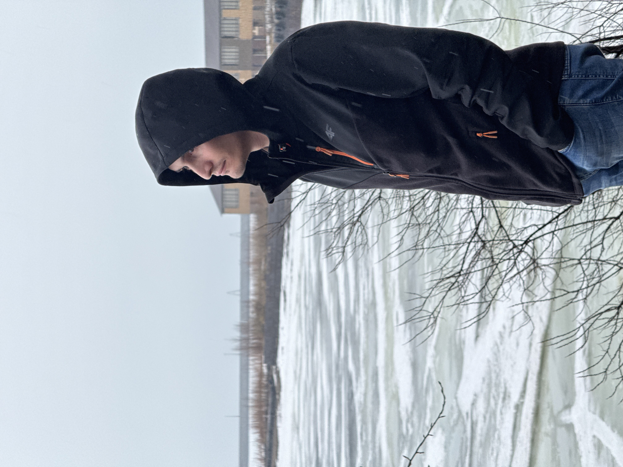 A young man in a black hooded jacket and blue jeans stands near what appears to be a frozen or icy body of water, possibly a river or lake. There are bare tree branches in the foreground. A multi-story building is visible in the background. The weather appears overcast, and it may be snowing lightly. The scene may be in or near the city of Chaikovsky, Russia.