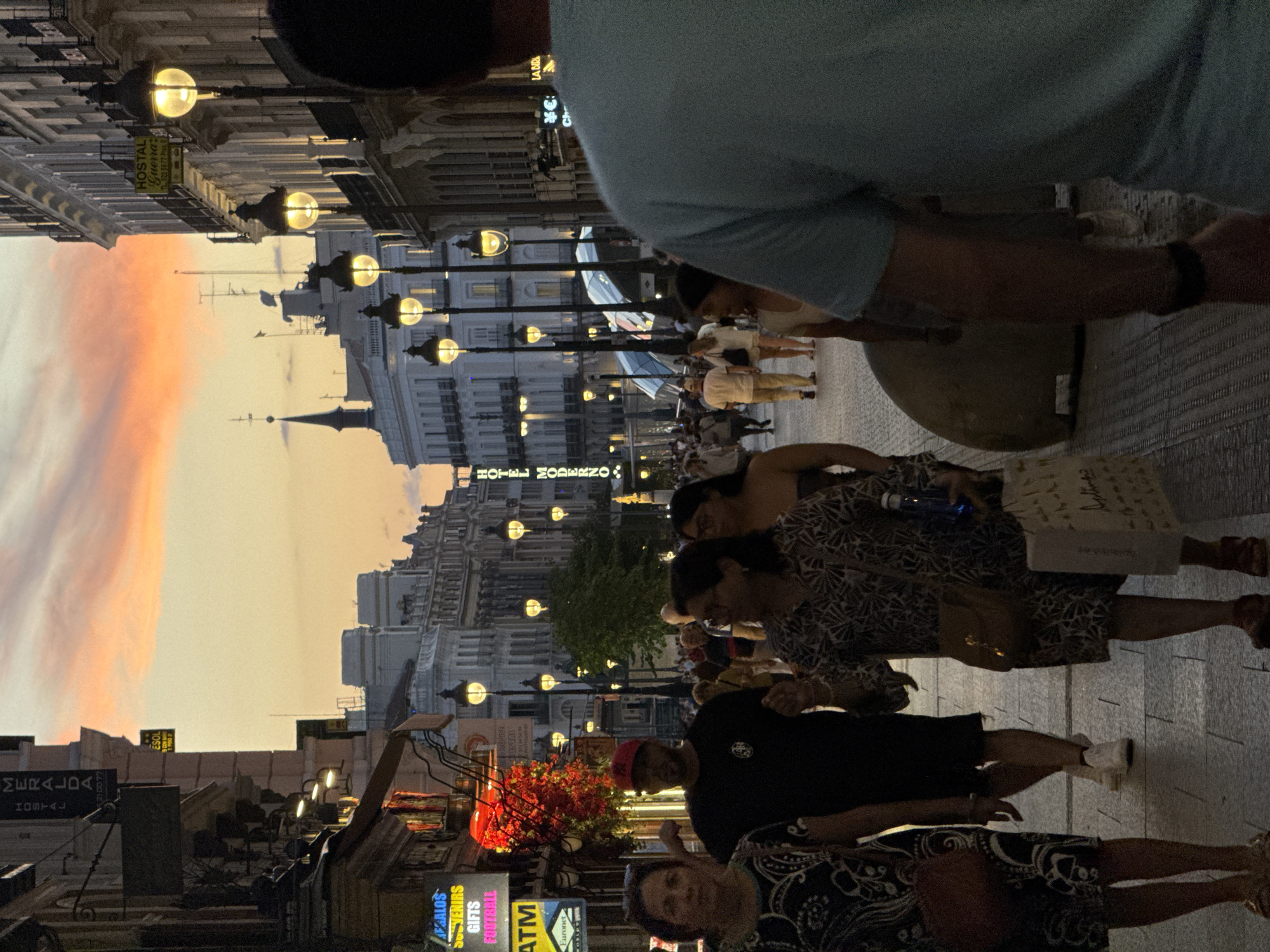 This photograph captures a bustling street scene in Madrid, Spain, likely during sunset or twilight. The sky exhibits hues of orange and pink, indicative of the setting sun. The street is lined with ornate, multi-story buildings, many featuring illuminated windows and balconies, suggesting they are shops, restaurants, or hotels. A prominent street lamp with multiple bulbs illuminates the pathway.
In the foreground, several people are walking along the cobblestone street. Closest to the camera, on the right, a person wearing a light blue t-shirt is partially visible from the back. To the left, a woman in a black and white patterned dress is walking, carrying a brown purse on her shoulder. Next to her, a man wearing a black t-shirt and a red cap is visible. Further back, other individuals and groups are walking, some are dressed casually, while others appear to be tourists. Shops with illuminated signs are visible on the left side of the street, including one advertising "ATM" and another with red flowering decorations. The overall atmosphere is one of urban activity and lively pedestrian traffic.
