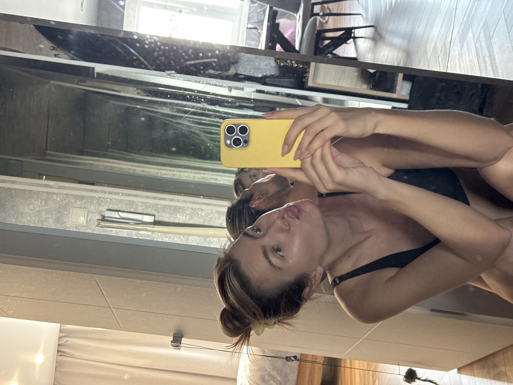 A young woman in a black undergarment is reflected in a mirror while holding a yellow smartphone. The mirror also reflects a room with a window and various items on a shelf. The scene is illuminated by natural light. The location of this scene in the city of Unknown, Unknown cannot be determined from the image.