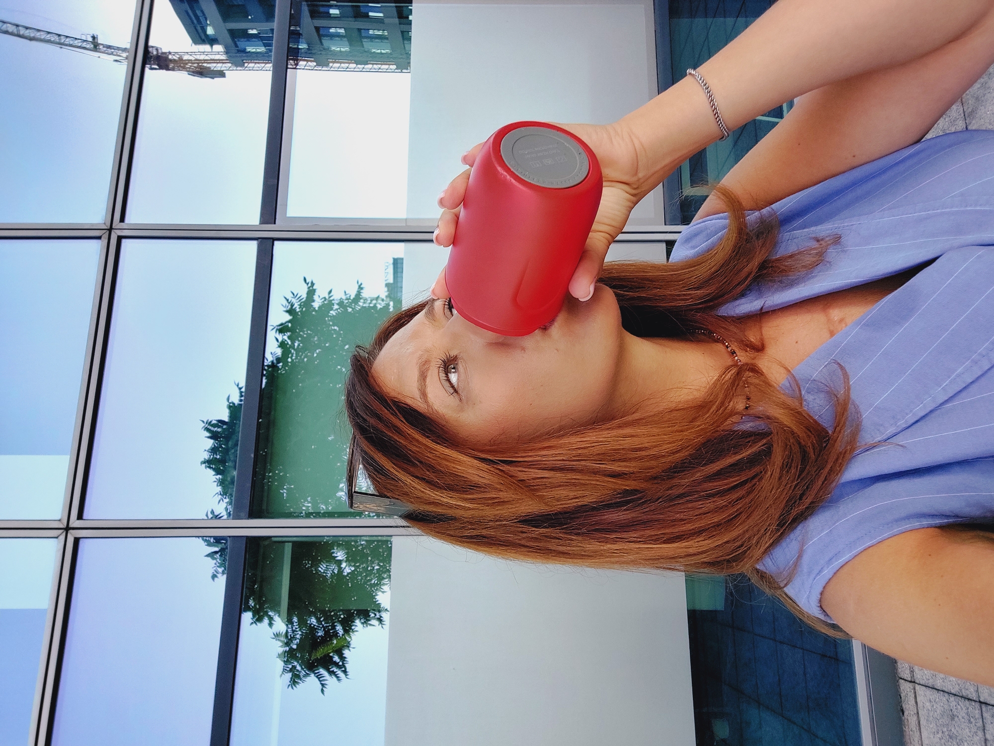 A woman with shoulder-length auburn hair is drinking from a red insulated tumbler. She is wearing a sleeveless periwinkle blue pinstripe blouse and a delicate gold necklace. Her right hand holds the tumbler to her lips. Her left arm is visible, adorned with a silver rope bracelet. She is positioned in front of a modern building with large, reflective windows. The windows reflect the sky, trees, and construction cranes, suggesting an urban setting in Sofia, Bulgaria. The lighting indicates it is daytime, possibly late afternoon, with soft light illuminating the scene.