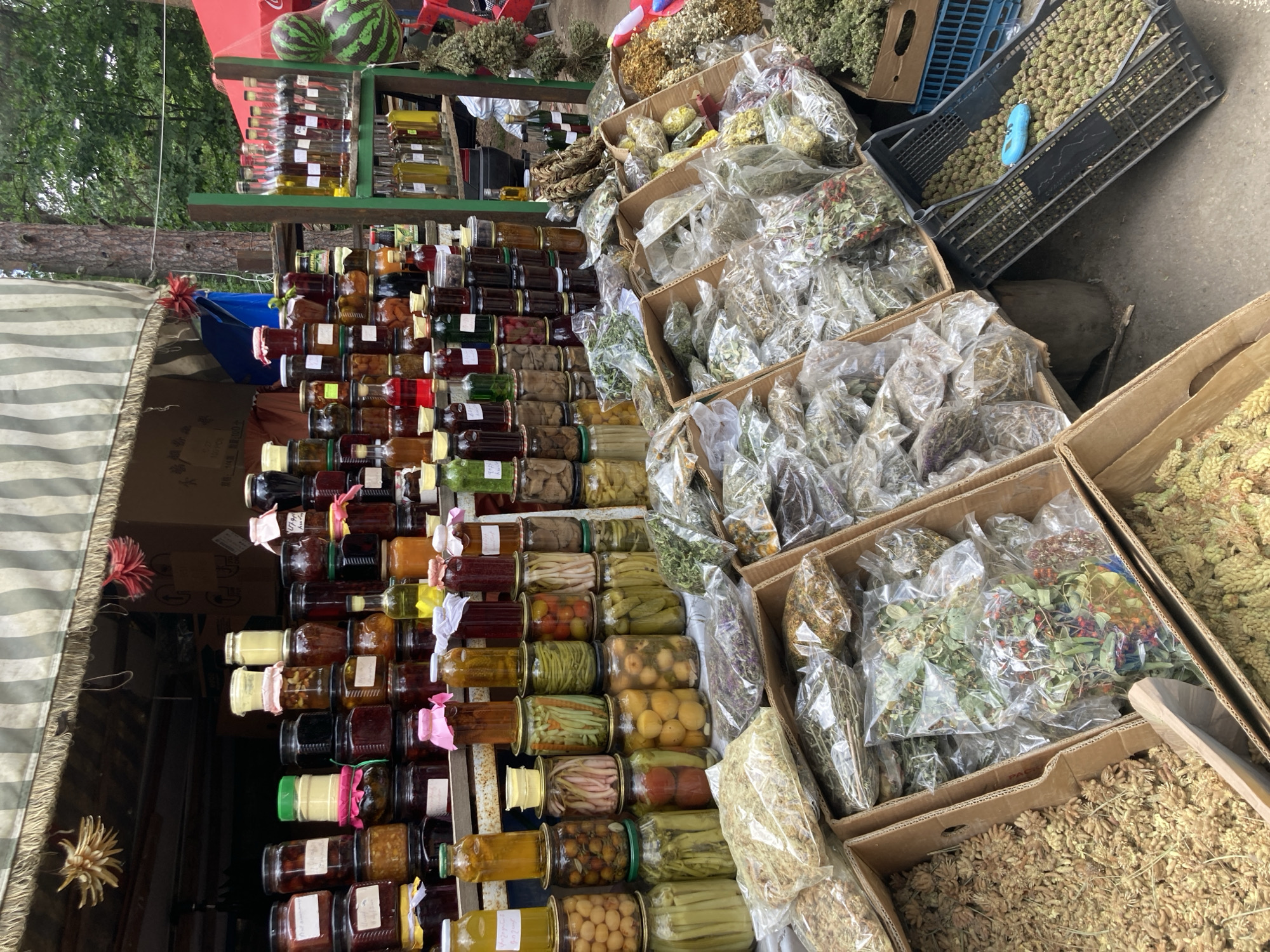 The image shows a market stall in Gyulagarak, Armenia, laden with various products. On the left, there's a tiered display of jars filled with preserves, jams, and pickled items. To the right are cardboard boxes and plastic crates containing dried herbs and herbal mixtures, some in clear plastic bags. Watermelons and bottles of liquids are visible on a higher shelf. A striped awning partially covers the left side of the stall.