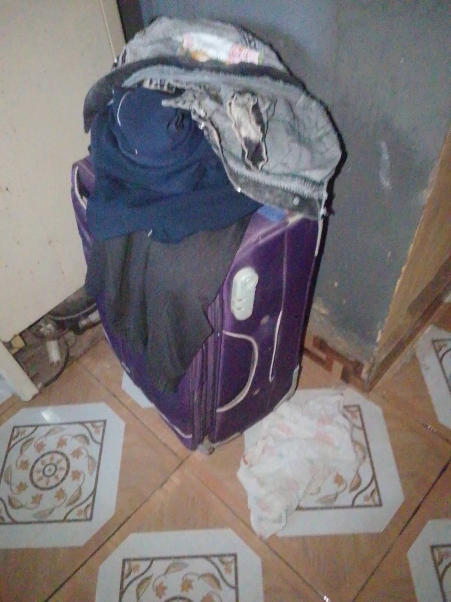 The image shows a purple suitcase overflowing with clothes. A grey jacket is draped over the top of the suitcase, and a dark blue garment is rolled up and placed inside. Next to the suitcase, on the floor, is a crumpled white piece of fabric with a faint floral pattern. The suitcase is positioned on a tiled floor with decorative square insets. In the background, a wall is visible, and part of a white appliance or cabinet. There is also some wiring and debris on the floor near the bottom left corner of the image. The lighting suggests it may be indoors. The scene appears to be a domestic setting, likely a room in Jalingo, Nigeria. No people are visible in the image.
