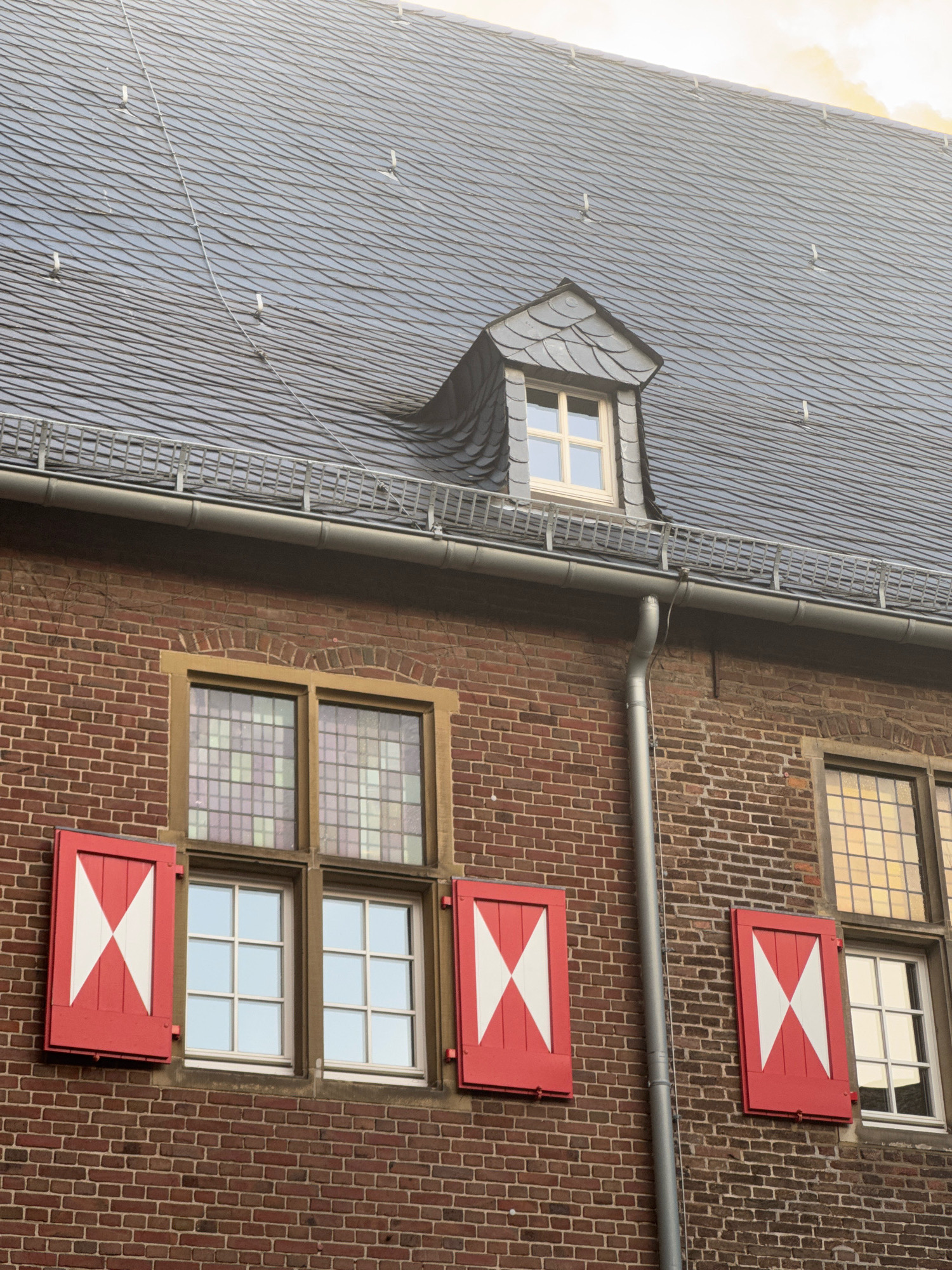 The image shows a close-up, angled view of a brick building with a dark slate roof. The building has several windows. Two windows are prominent on the lower left, each with a wooden frame and a grid pattern of smaller panes. Red shutters with white 'X' designs are closed over the lower halves of these windows. To the right of these, another window is visible with a similar grid pattern. Higher up on the roof, a small dormer window with a light-colored frame and grid is visible. A metal downspout runs vertically along the brick wall. The overall impression is of historic architecture. The location is identified as Cleves, Germany.