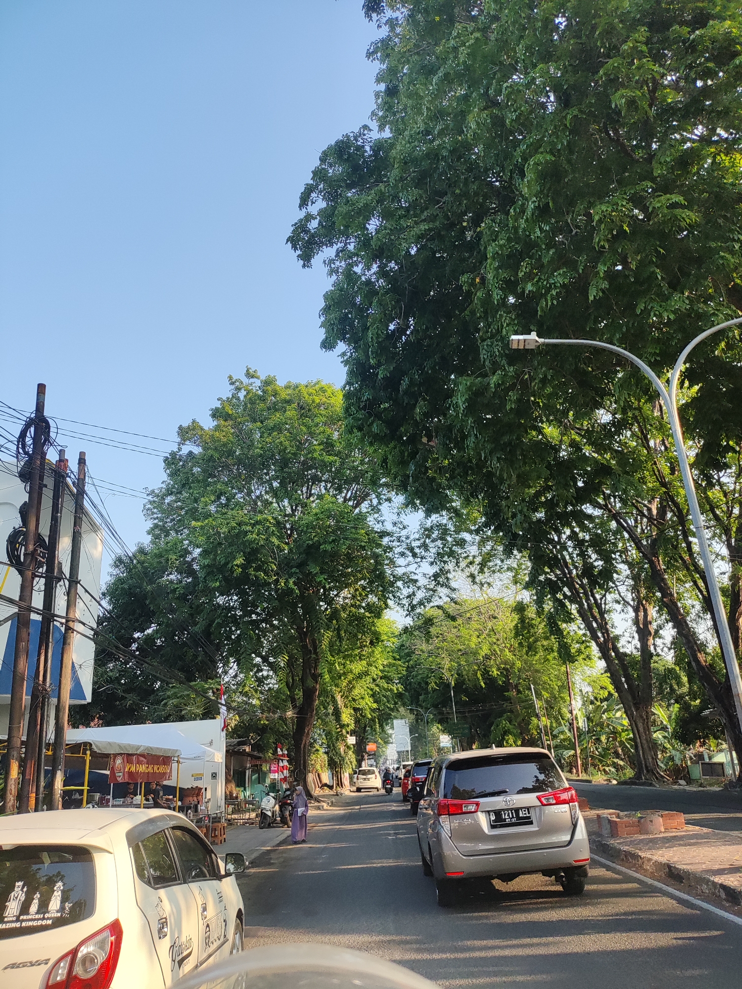 The image depicts a street scene in Cirebon, Indonesia, under a clear blue sky. The foreground is dominated by the rear of a white car, on which several stickers are visible, including one that reads "KING PRINCESS QUEEN AMAZING KINGDOM". To the left, there is a white building with awnings and yellow poles, suggesting a commercial establishment, possibly a food stall, with people visible inside and nearby. A woman in a purple hijab is walking on the sidewalk. In the middle ground, a silver SUV with license plate "D 1211 AEL" is driving on the road, followed by other vehicles. The street is lined with mature trees casting shadows on the asphalt. Utility poles with numerous wires are also present on the left side of the road. The overall scene suggests a typical urban environment during daylight hours.