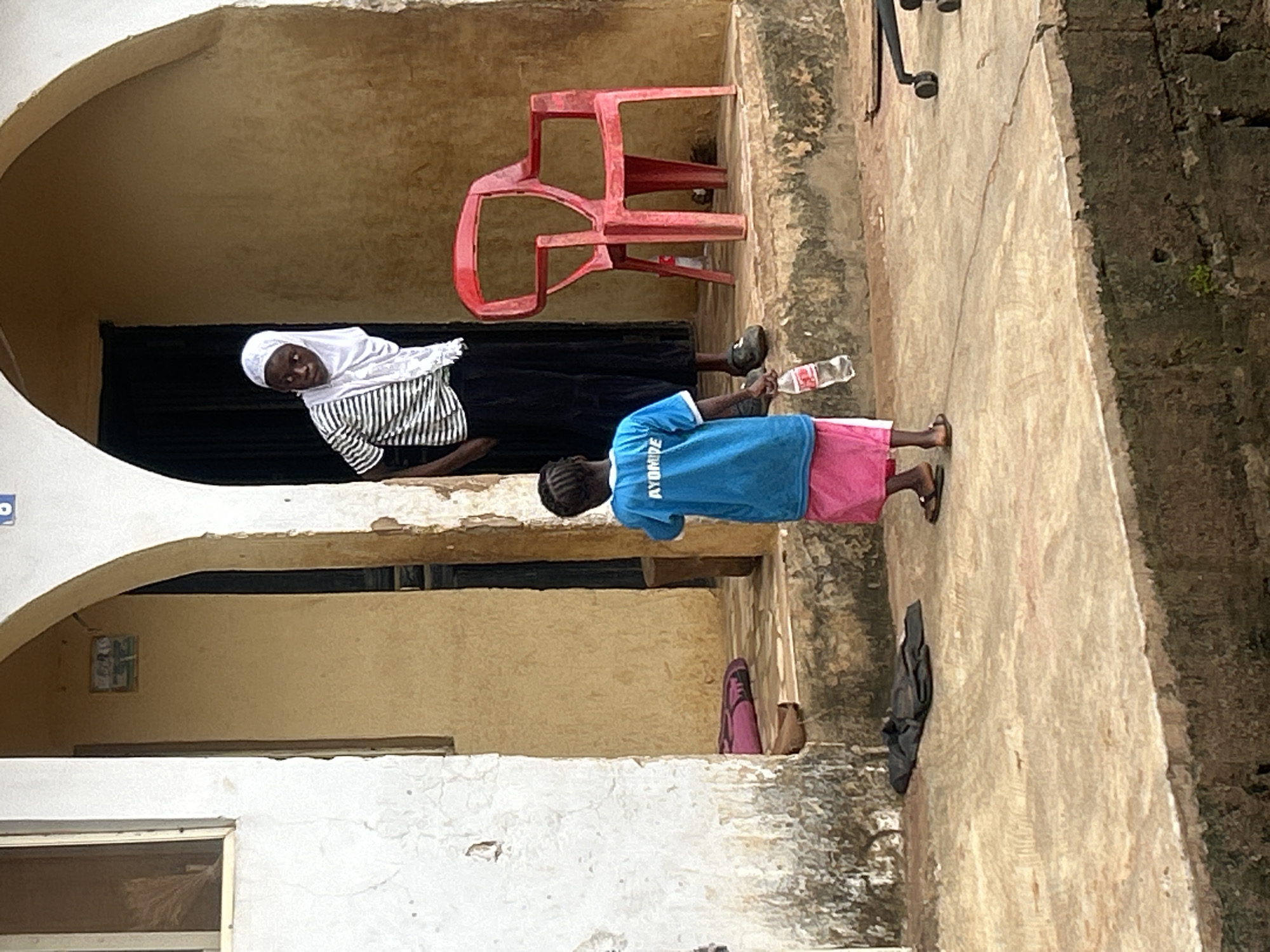 An outdoor scene depicts two females in Ikorodu, Nigeria. The foreground shows a young female wearing a blue t-shirt with the inscription "AYOMIDE" and a pink skirt, holding a plastic water bottle. She is standing on a concrete surface with a few dark clothes on the ground behind her. To her right, an adult female wearing a white hijab and a striped t-shirt over a black skirt stands near a bright red plastic chair. The background features an arched entrance to a building with yellow walls, and part of a white exterior wall. The environment appears to be a residential area with a slightly weathered appearance.