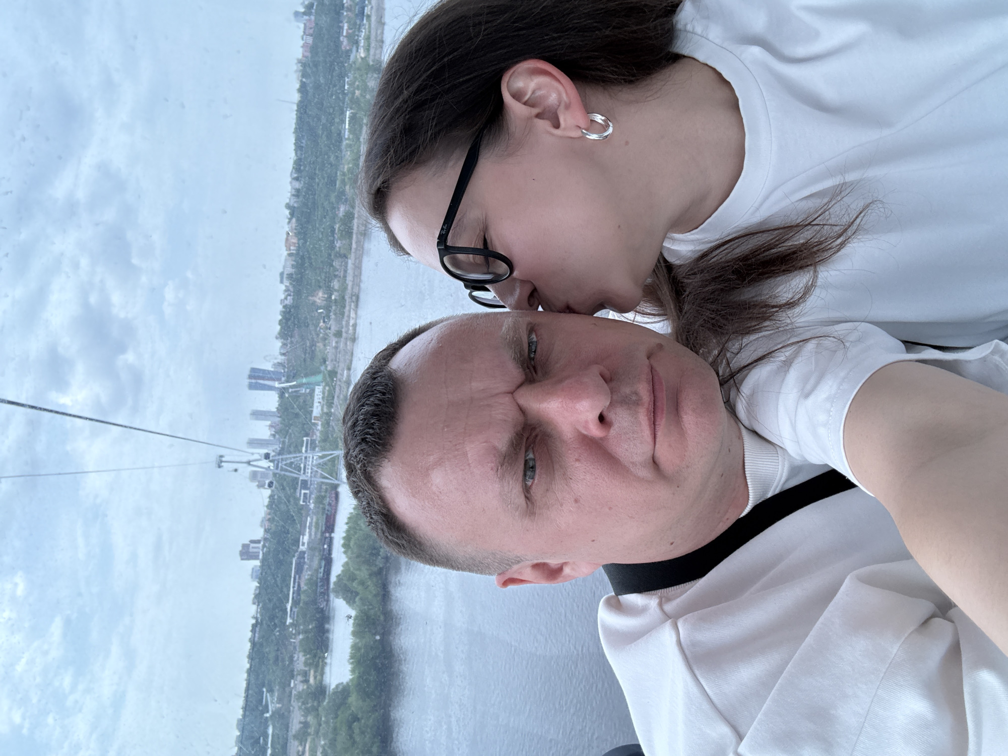 A selfie taken from inside a cable car. Two people are visible: an adult man and a younger adult woman. The woman is positioned slightly above and behind the man, and she appears to be kissing his forehead. Both individuals are wearing white T-shirts. The woman has dark hair, wears glasses and a hoop earring. The man has short, graying hair. The background shows a cityscape through the cable car window, which appears to be the city of Bor, Russia, based on the visible cable car infrastructure and city architecture. A river is visible below, with the cityscape extending along the opposite bank. The sky is overcast.