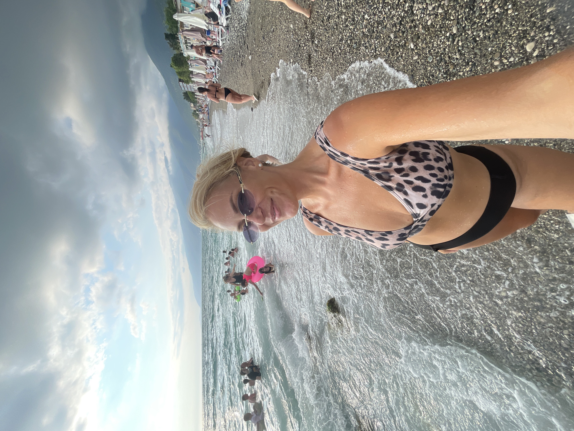 A woman in a leopard print bikini and black bottoms is taking a selfie on a pebble beach in Gagra, Abkhazia. She is wearing oval sunglasses. The water is shallow, with waves lapping at her feet. Several other people are visible in the ocean, some swimming and others with inflatable toys. In the background, the beach is lined with umbrellas and lounge chairs, with buildings and mountains further beyond. The sky is partly cloudy.