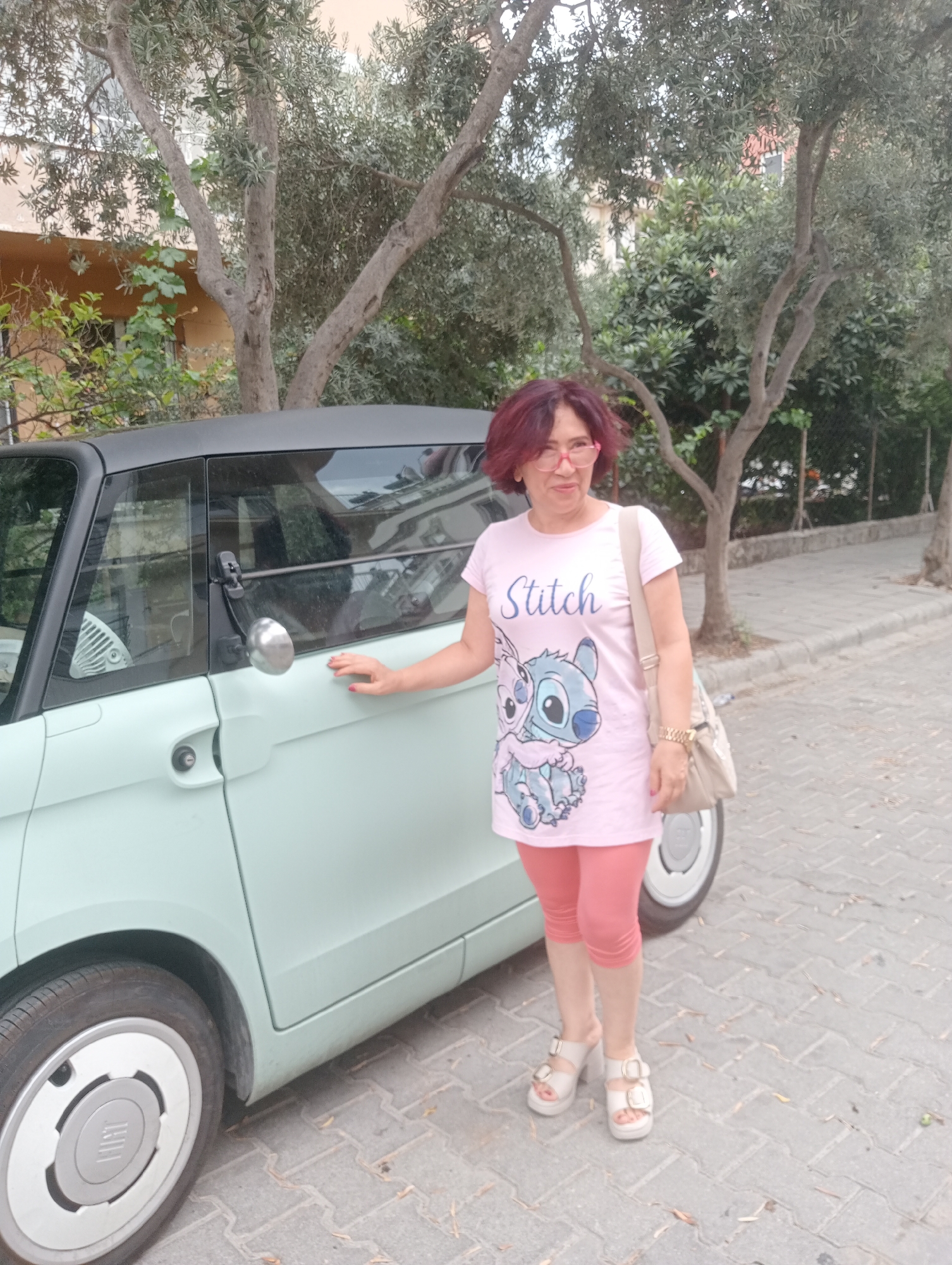 An adult woman stands next to a light mint green car on a paved street in Fethiye, Turkey. The woman has short, dark red hair and wears pink-rimmed glasses, a light pink t-shirt with a Stitch graphic and the word "Stitch" printed on it, and coral-colored capri leggings. She has on light-colored platform sandals. A beige shoulder bag is slung over her right shoulder. Her left hand is resting on the car door. The car appears to be a small, modern vehicle with a black roof and a silver side mirror. The background features trees with olive-like leaves, a building with a light brown facade, and a sidewalk. The ground is paved with gray interlocking bricks. Fallen leaves are scattered on the ground. The scene is outdoors and appears to be daytime.