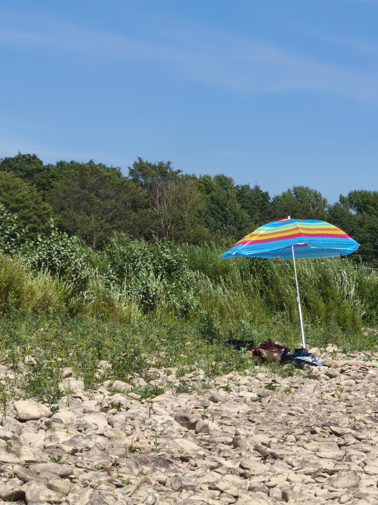 An outdoor scene features a beach umbrella on a rocky shore. The umbrella is multicolored with stripes of blue, yellow, pink, and orange. It is anchored in the ground by a white pole. In the background, a dense line of green trees forms a backdrop against a clear blue sky. On the ground near the umbrella, several items are present, including a brown bag, a red garment, and a folded blue and white striped cloth. The shore is covered in light-colored rocks of various sizes.