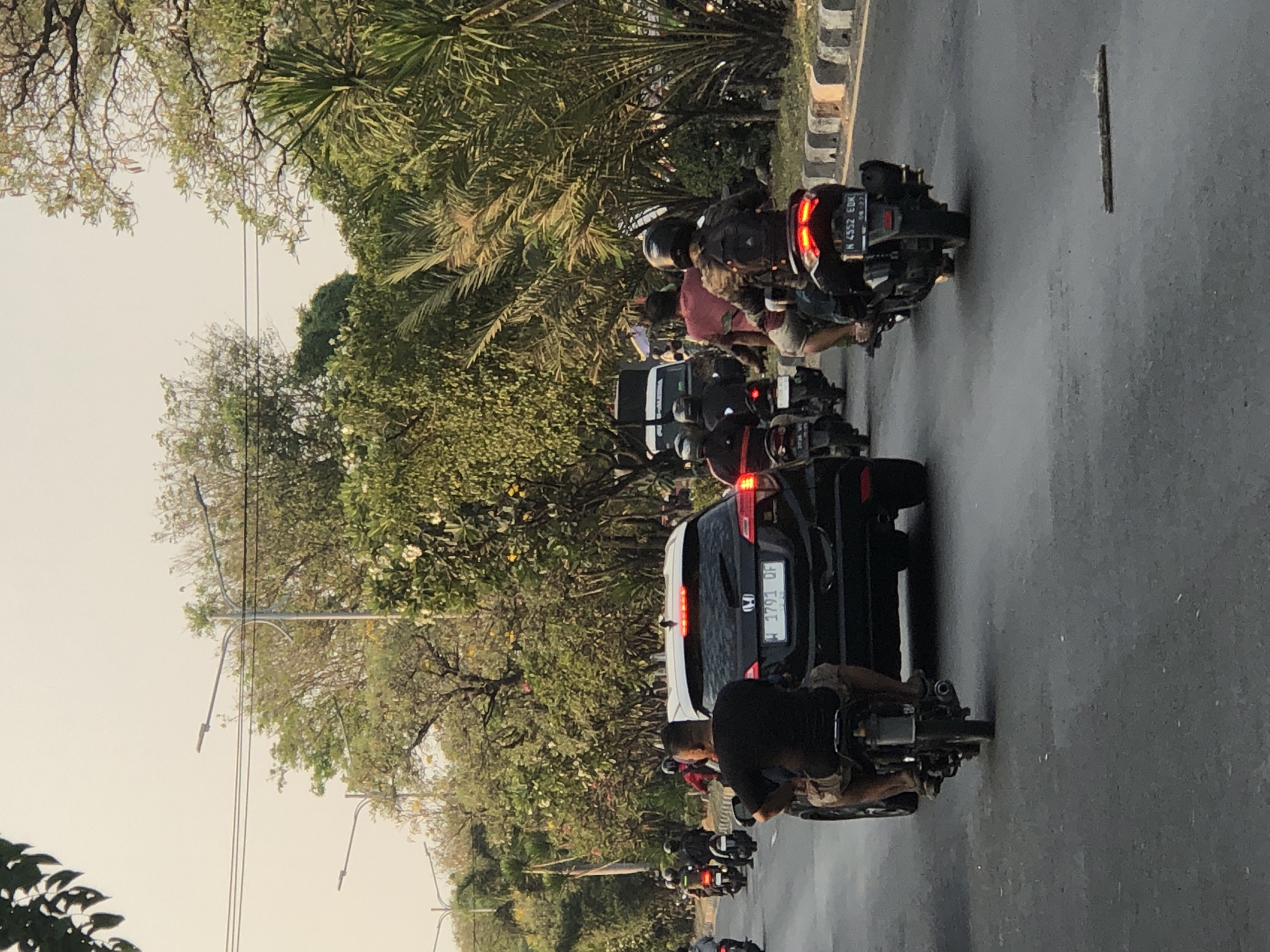 The image shows a traffic scene on a road in Ngaban, Indonesia. Several motorcycles and a black SUV are visible. A person in a black t-shirt and shorts is riding a motorcycle in the foreground, facing away from the camera. Behind them, the black SUV is also moving forward. On the right side of the road, more motorcycles are traveling in the same direction. The road is bordered by a sidewalk with a concrete barrier and lined with trees and foliage. Streetlights are present along the side of the road. The sky is bright, suggesting it is daytime, possibly late afternoon or early evening due to the lighting.