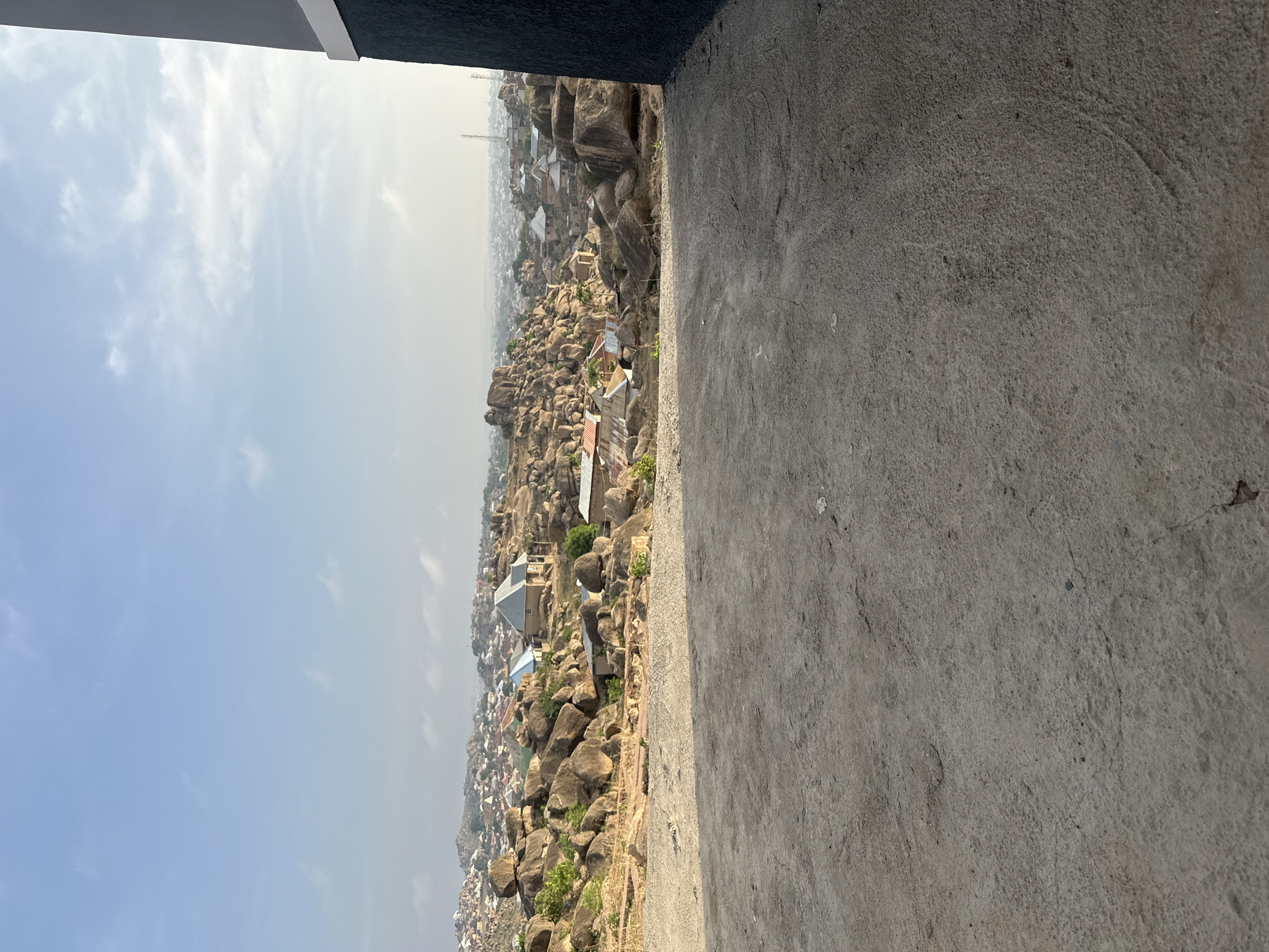 The image shows an elevated view of a landscape, likely within the city of Jos, Nigeria, known for its rocky terrain and dense urban development among the rock formations. The foreground features a concrete surface of a building or structure, suggesting the perspective is from a rooftop or high vantage point. The middle ground consists of a dense concentration of rock formations interspersed with residential buildings, exhibiting a mix of traditional and modern architectural styles, characterized by red and blue roof colors. The background reveals a hazy sky with limited visibility, indicating possible air pollution or dust. No people are directly visible in the scene. The overall setting conveys an urban landscape integrated within a natural, rocky environment.