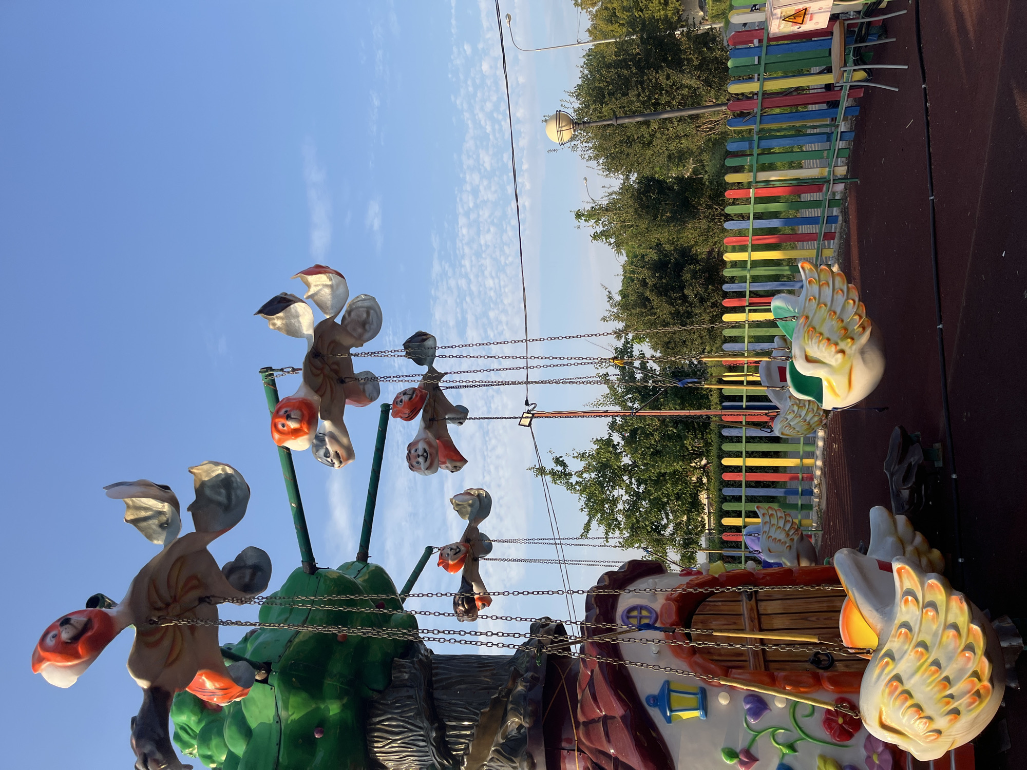 An amusement park ride, designed as a whimsical treehouse with hanging swings shaped like colorful cartoon animals, is captured under a bright blue sky with scattered clouds. The ride features a central tree structure with a fairytale-like house at its base, adorned with painted flowers and a wooden door. Suspended from the branches of the tree are chains holding individual swings. Some swings are designed as cartoon animal heads, possibly lions, with orange and white faces. Others are shaped like large, stylized swan or duck-like figures with feathered wings painted in shades of white, yellow, and orange. The ride is surrounded by a colorful, striped wooden fence. In the background, lush green trees are visible, along with streetlights and utility poles. The ground beneath the ride appears to be a dark, rubberized surface, common in playgrounds. A small, three-legged stool with a light-colored seat stands near the fence, next to an electrical box with a yellow warning symbol. The overall scene suggests a public recreational space, likely a park or a small amusement area within a city. The image does not contain any identifiable people.