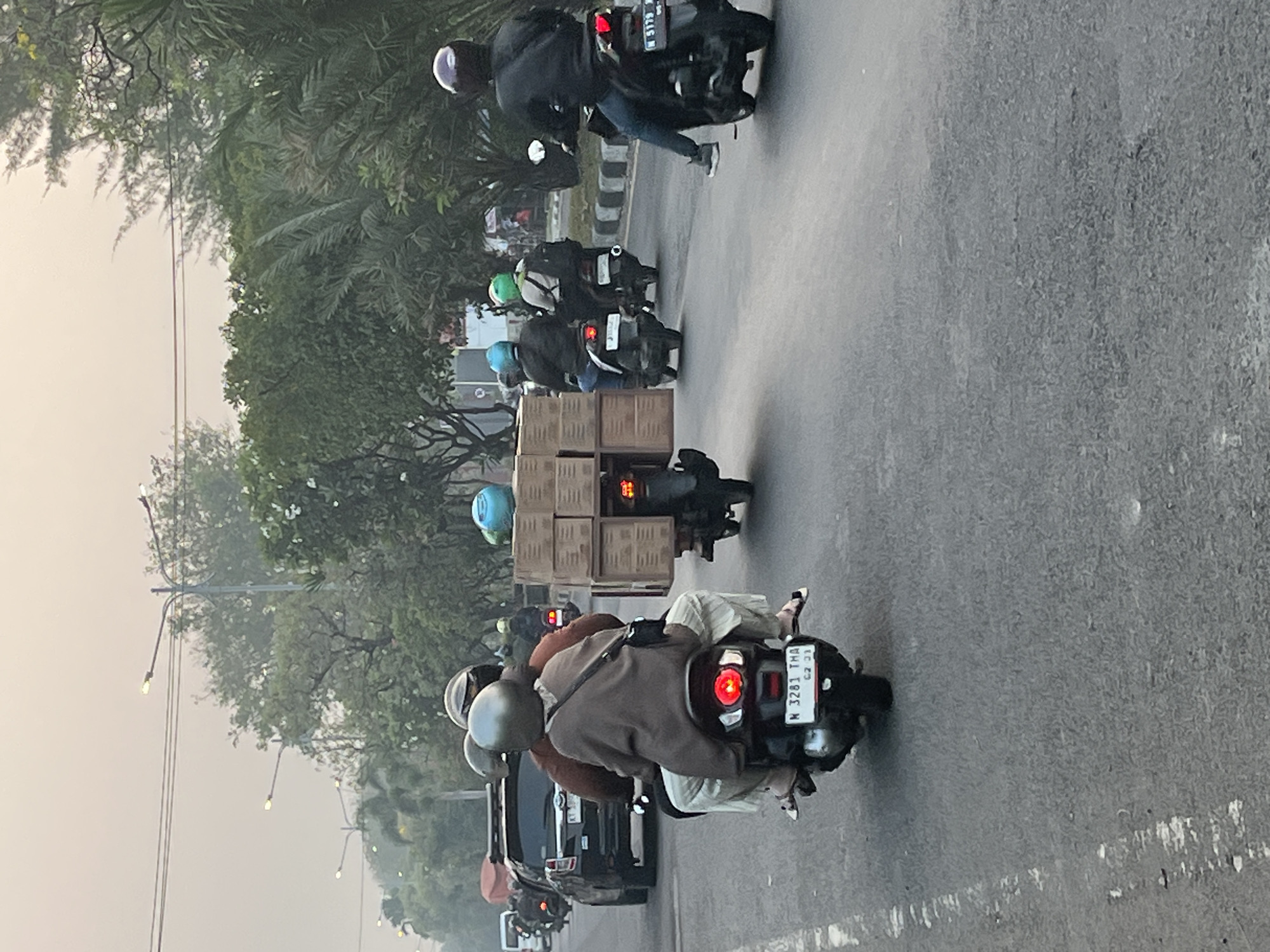 The image depicts a street scene in Kludan, Indonesia, with several motorcycles navigating traffic. In the foreground, a motorcycle carries two passengers, a driver and a passenger, both wearing helmets. The passenger in the back is wearing a brown overshirt and light-colored pants, and is looking forward. The motorcycle has a license plate that reads "N 3281 THA CZ 03". To the right of this motorcycle, another motorcycle is carrying a stack of cardboard boxes, also with two passengers wearing helmets. Further down the road, more motorcycles are visible, some carrying additional passengers or cargo. A black SUV is seen in the left lane. The environment appears to be a moderately busy street with trees and streetlights lining the roadside. The sky is overcast, suggesting either early morning or late afternoon.