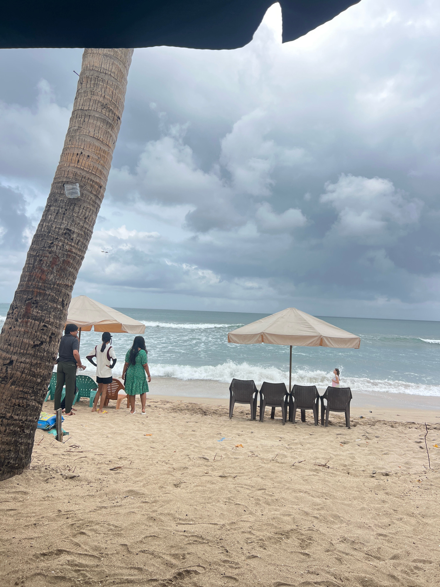A cloudy day at Kuta Beach, Indonesia, is depicted with a focus on the sandy shore and the ocean. In the foreground, a thick, textured palm tree trunk dominates the left side of the frame, with a small white object attached to it. Three people are visible near the palm tree, facing the ocean: two standing and one slightly behind them.
Further down the beach, two beach umbrellas are set up. Under the first umbrella, which is a light beige color, there are three plastic chairs arranged. To the left of this umbrella, another person is standing. Under a second, larger umbrella to the right, four dark plastic chairs are arranged in a row.
The ocean is active with moderate waves breaking on the shore. A single person, wearing a pink top, is visible in the shallow water near the second umbrella. The sky is filled with heavy, gray clouds, suggesting overcast or impending rain. The sand appears dry and textured with scattered debris like small sticks and leaves. The overall scene conveys a slightly subdued atmosphere due to the cloudy weather.