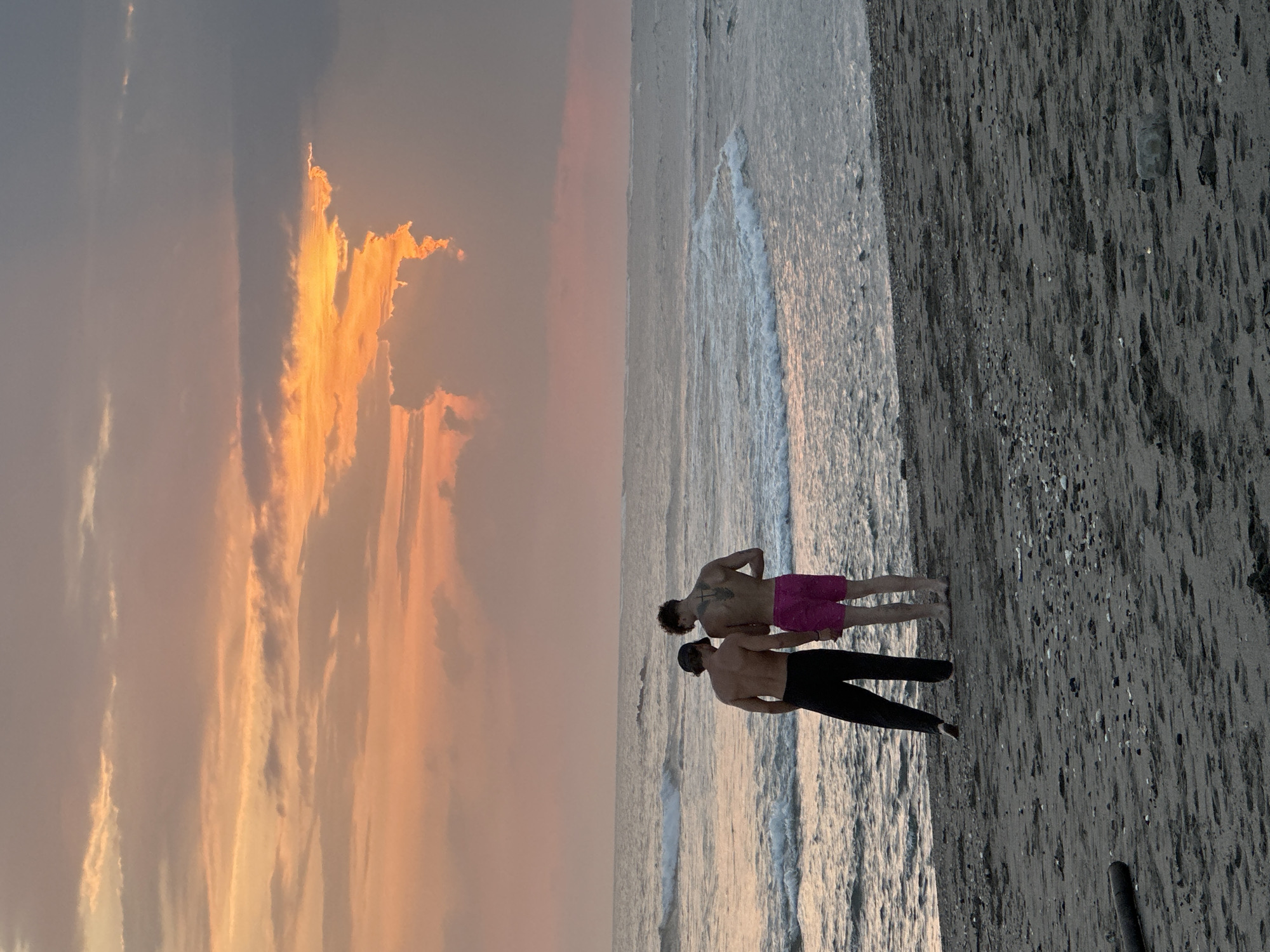 Two shirtless young men stand on a sandy beach, facing the ocean, during sunset. The man on the left wears black pants and a dark baseball cap. The man on the right wears bright pink shorts and has a large tattoo on his back. Waves gently break on the shore, and the sky is a mix of orange, pink, and gray clouds, illuminated by the setting sun. The beach is dark sand with scattered pebbles and debris. The scene is likely located on the coast of Mengwi, Indonesia, a region known for its beaches and sunsets.