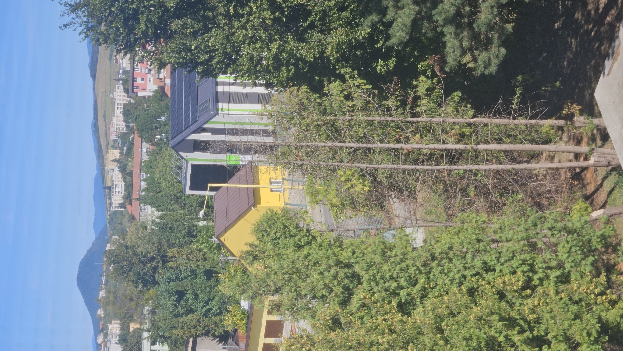 The image is a distant view of a residential area in Prešov, Slovakia, under a clear blue sky. In the foreground, several evergreen trees are visible, with some appearing dry and bare. Behind them, a bright yellow house with a brown roof stands next to a modern, two-story structure with green and white striped siding and solar panels on its roof. A yellow lamppost is also present near the yellow house. In the mid-ground, a network of trees separates the foreground from the city. The background reveals a densely populated urban landscape with various apartment buildings and houses, some with red roofs. Further in the distance, rolling hills and mountains form the horizon, with a prominent, cone-shaped hill in the left background that appears to have a structure on its peak. The overall scene depicts a sunny day in a suburban environment that blends with the urban sprawl of Prešov.
