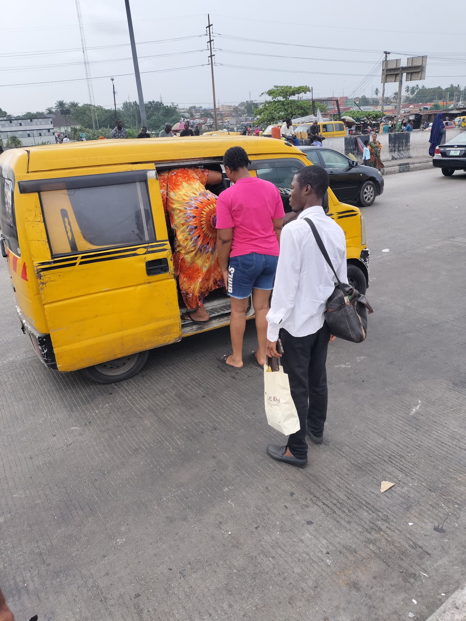 In Ikeja, Nigeria, a yellow minibus, likely a public transport vehicle known locally as a "danfo," is parked on a gray asphalt surface. A woman in a vibrant orange and yellow patterned dress is entering the bus through the open sliding door. A second woman, wearing a pink shirt and blue denim shorts, is positioned behind her, also about to board. Beside the bus, a man in a white shirt and black pants stands with a black shoulder bag and a white shopping bag with text written on it. Other cars and people can be seen in the background indicating a busy urban environment. Power lines are visible overhead.