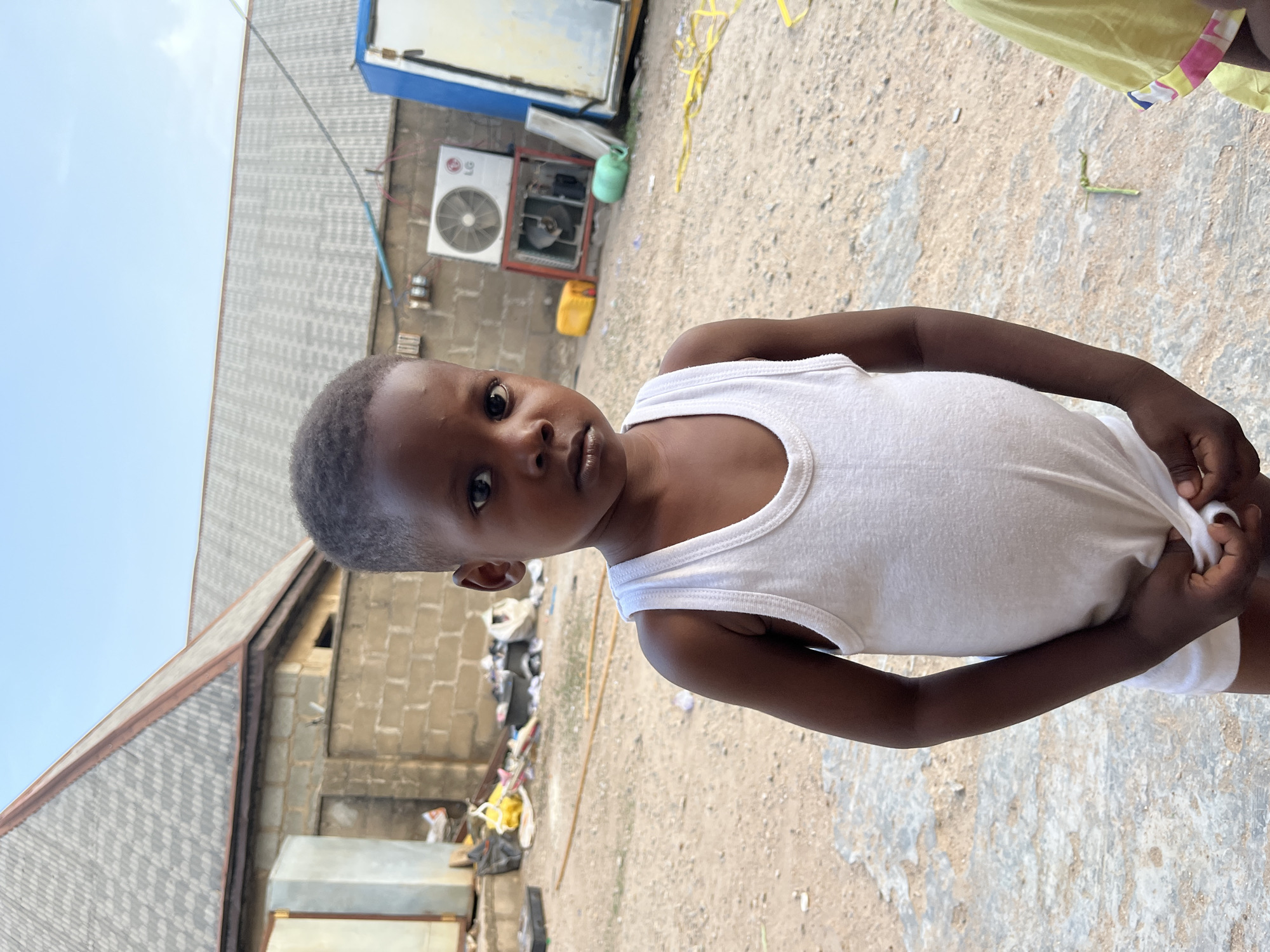 A young child wearing a white undershirt stands outside in Iddah, Nigeria. The location appears to be near or behind buildings made of concrete blocks. There is debris scattered on the ground, and an air conditioning unit is visible on one of the buildings. The child is looking toward the camera. The sky is visible in the top left of the frame.