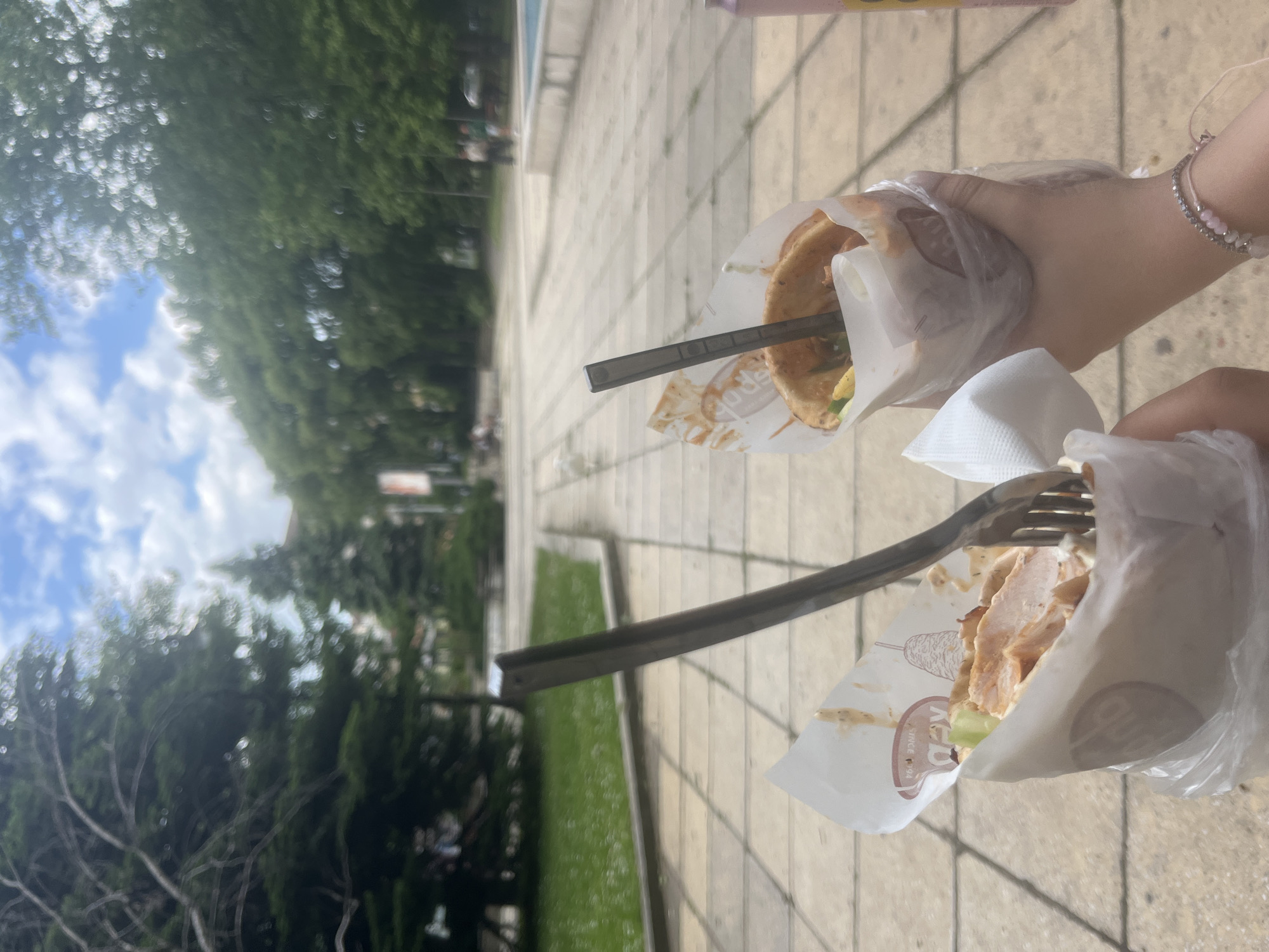 The image depicts a ground-level view outdoors, likely on a sidewalk or pedestrian area in Varna, Bulgaria. Two hands hold food items, each pierced with a fork. The food appears to be gyros or similar sandwiches in paper wrappers and plastic bags. The surroundings include paved ground, and some vegetation in the background, including grass and trees. The sky is visible through the trees, with a mix of blue sky and clouds. The overall scene suggests an informal outdoor setting, possibly near a park or in a tourist area. The light is bright, indicating a sunny day.