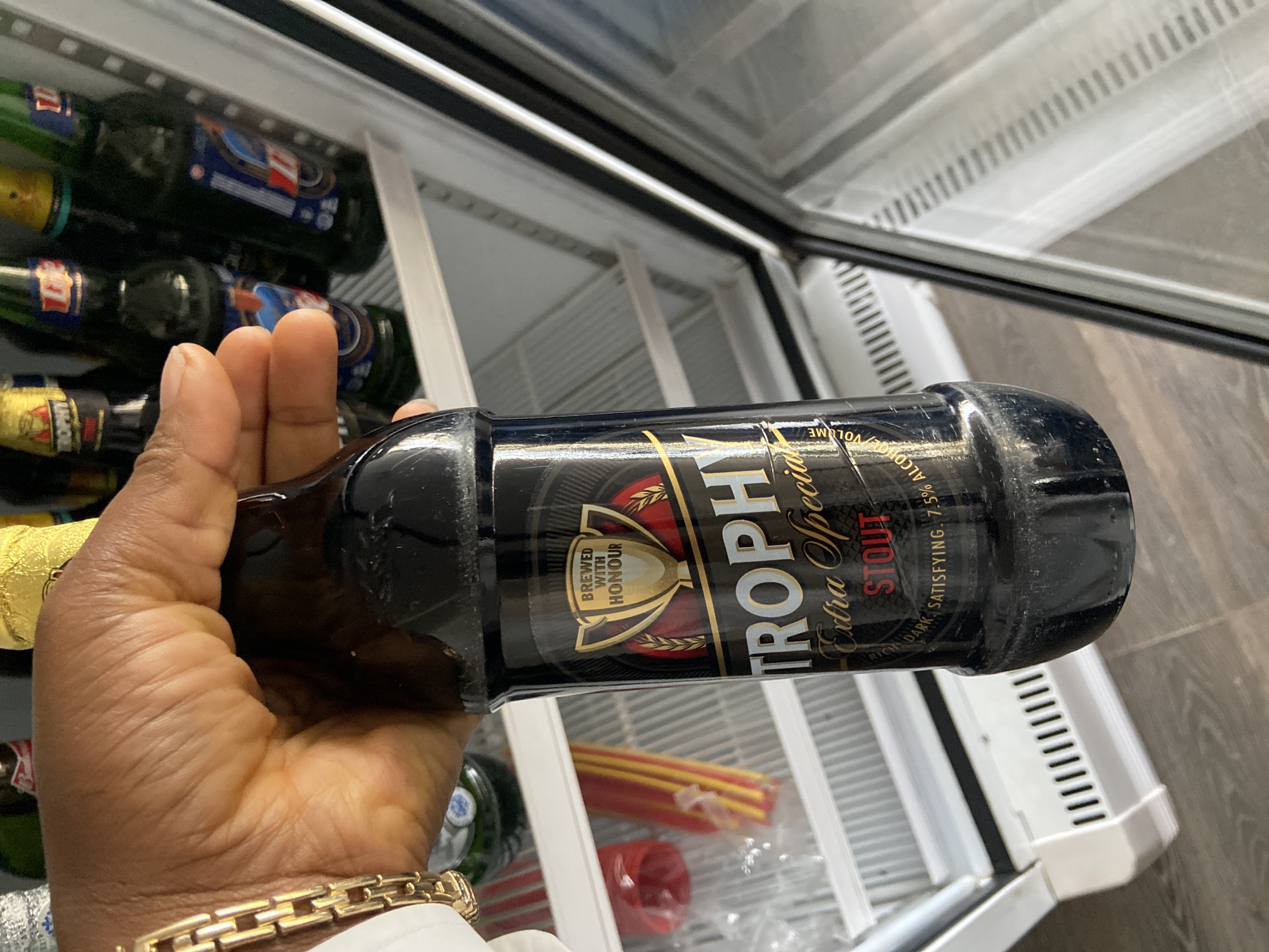 A person's hand, adorned with a gold bracelet, holds a dark glass bottle of "Trophy Extra Special Stout" in front of an open, white-interior refrigerator. The bottle's label is predominantly black with gold lettering and features a golden trophy emblem. The refrigerator shelves are visible behind the bottle, containing other bottles and some red and yellow striped items. The scene appears to be indoors, possibly in a home or a commercial establishment in Gura Top, Nigeria, given the branding of the beer. The lighting suggests an indoor environment, with some reflections on the refrigerator door.