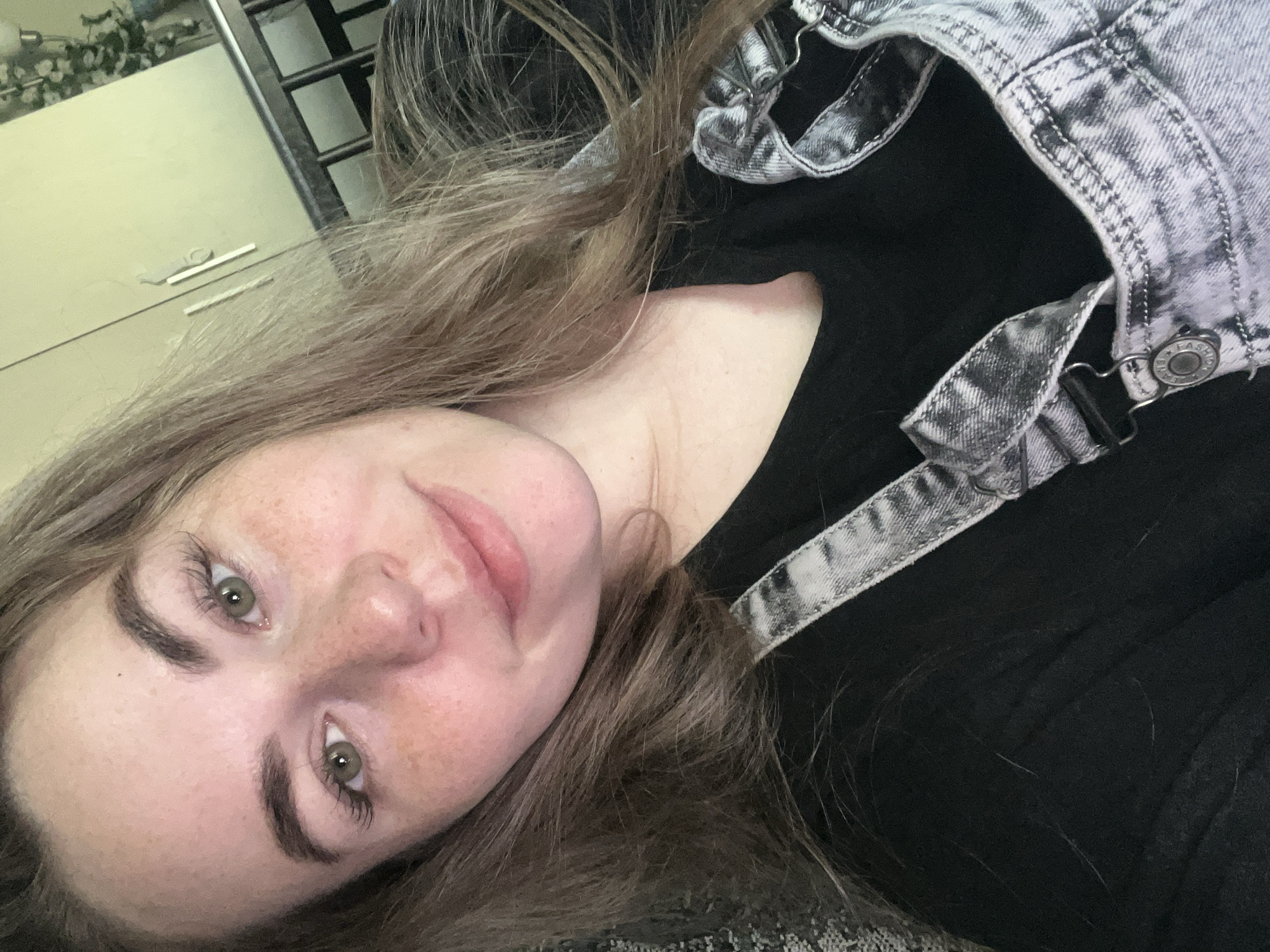 The image is a selfie of a young woman. She has fair skin with freckles, light brown hair, and green eyes. She's wearing a black top under a pair of light gray denim overalls. The background includes a glimpse of a light-colored wall and a metal structure, possibly part of a bed frame or shelf. There is no information indicating that the image was taken in Chaikovsky, Russia.