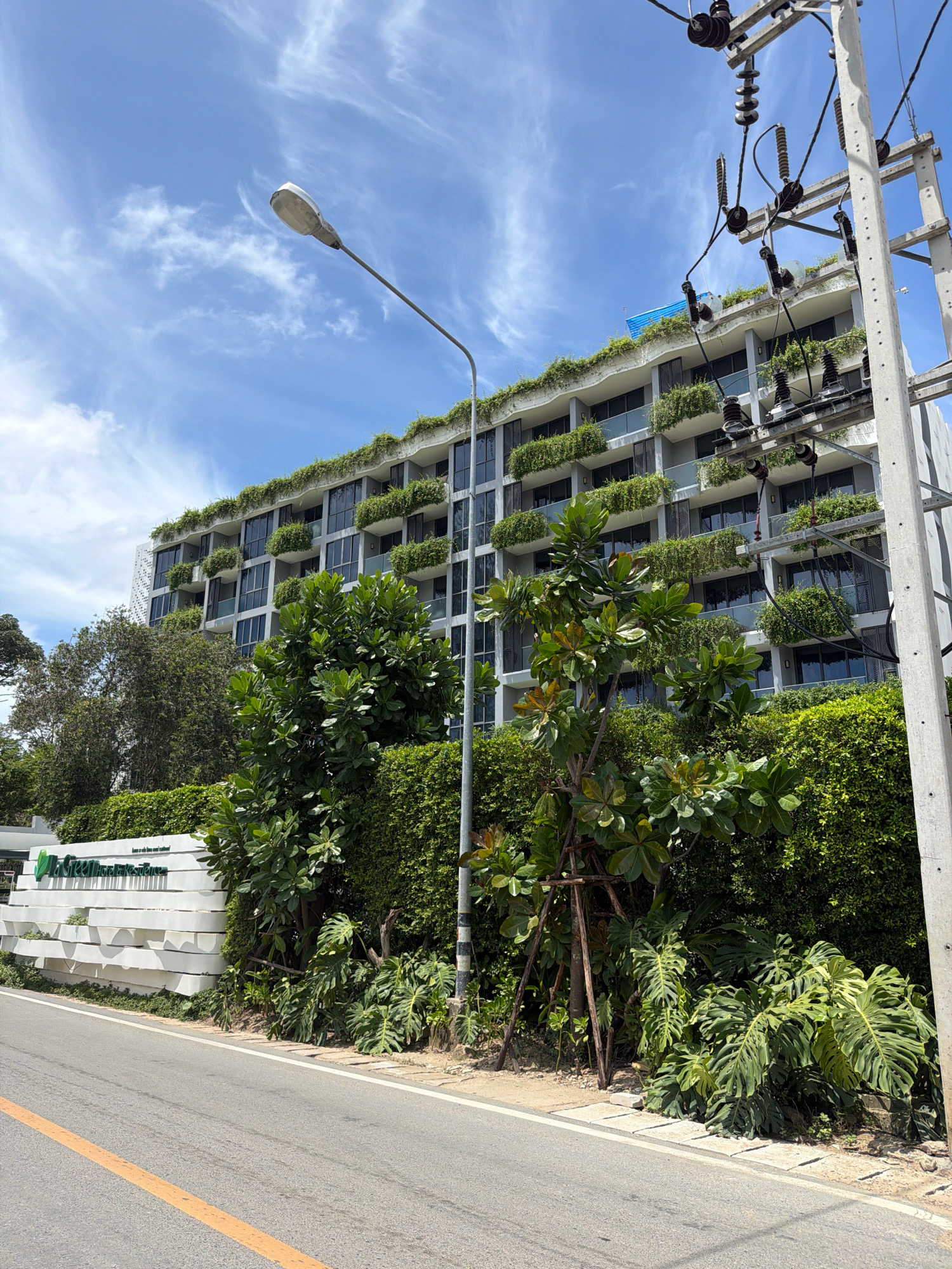 The image shows a modern, multi-story building in Choeng Thale, Thailand, with a significant amount of greenery on its balconies and rooftop. The building appears to be a residential complex or hotel, with numerous windows and balconies visible. In the foreground, there is a paved road with a yellow line marking the lane. To the left of the road, there is a white wall with the text "La Green Garden Residences" written in green. This wall is adorned with a modern, stepped design and is surrounded by lush vegetation, including trees and bushes. On the right side of the road, a utility pole with electrical wires and insulators is visible. The sky is blue with scattered clouds. The overall impression is one of a well-maintained property integrated with nature.