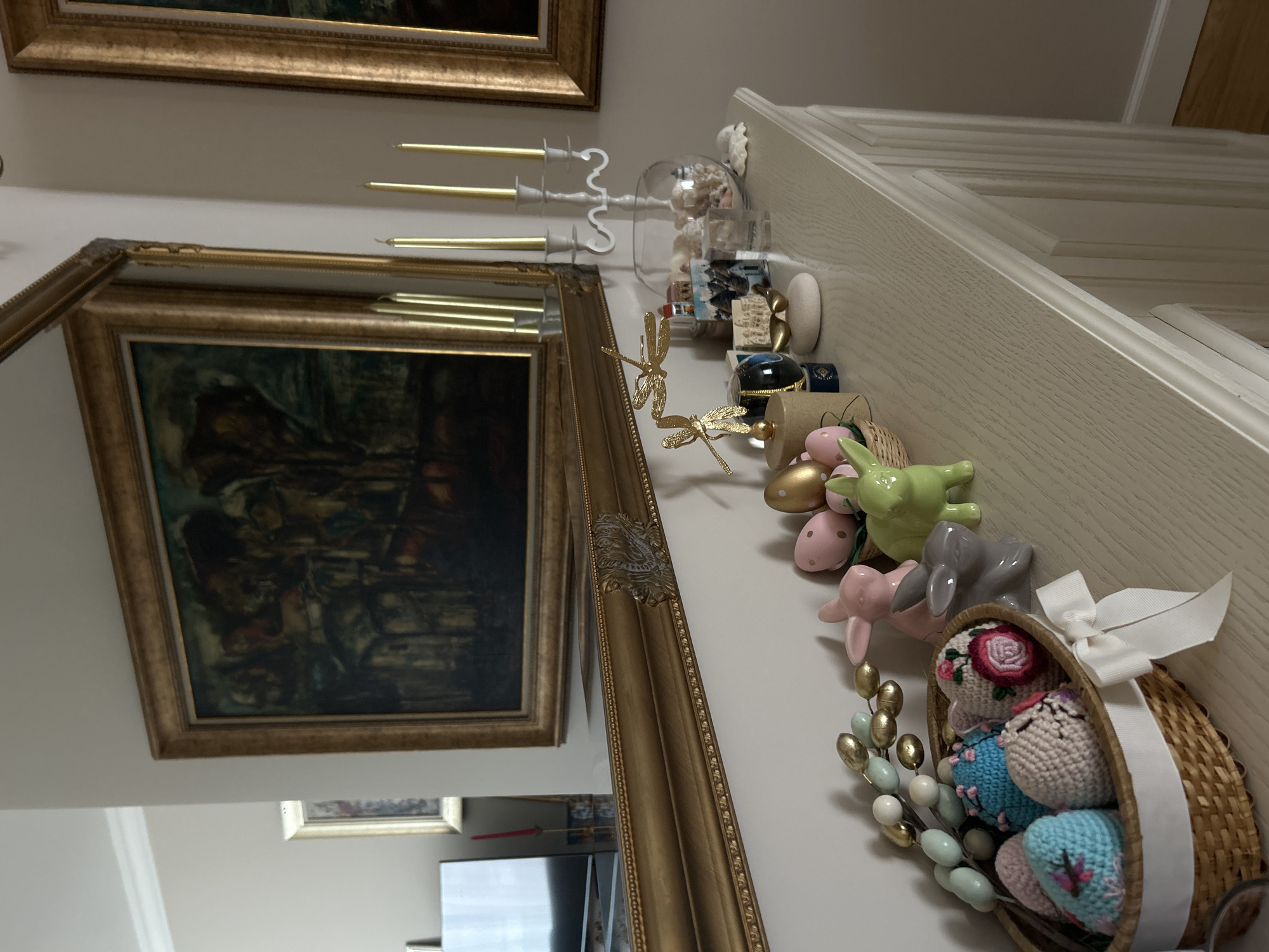 The image shows a wall in a room, potentially in a residential setting, decorated with various items. A large, ornate gold-framed mirror and a similarly styled painting hang on the wall. A collection of decorative objects, including ceramic bunnies, golden eggs, and a small woven basket filled with fabric eggs, sits on a ledge or shelf running along the wall. A three-armed candlestick holder with gold candles is also visible. The reflection in the mirror shows portions of a room interior, including a television screen and other decorative items, potentially indicating the location in Plovdiv, Bulgaria, is within a home.