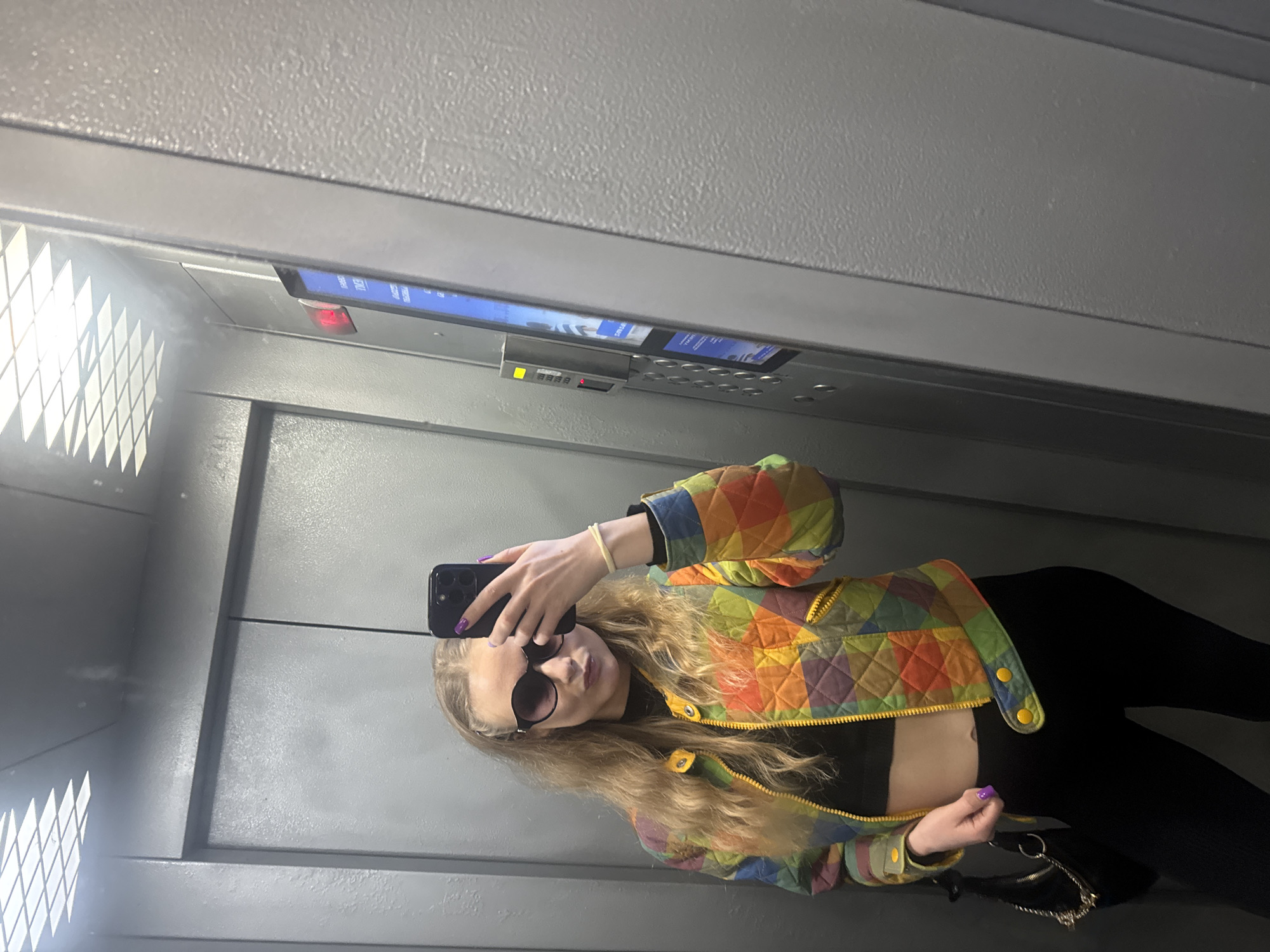 A young woman is taking a selfie in an elevator in Unknown, Unknown. She is wearing a colorful quilted jacket, black leggings, and sunglasses. She is holding a black smartphone in her left hand. The elevator has a gray interior.
