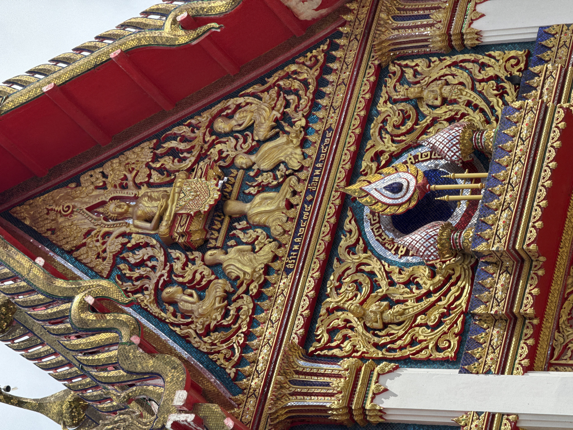 This is a close-up, eye-level shot of the exterior facade of a temple in Chalong, Thailand. The architecture is highly ornate, featuring intricate gold detailing and vibrant red roof elements. At the apex of the gable, a golden statue of Buddha is seated. Below this, a row of smaller golden figures, possibly monks or deities, are depicted in various postures. Decorative scrollwork, also in gold, adorns the facade. A band of Thai script is visible across the upper section of the facade, beneath the row of figures. Below this, a decorative archway frames a central ornament with a teardrop shape, surrounded by patterned tiles and small golden pillars. The entire structure appears to be adorned with mosaic tiles in shades of blue, red, and gold. The sky is a pale, overcast white.
