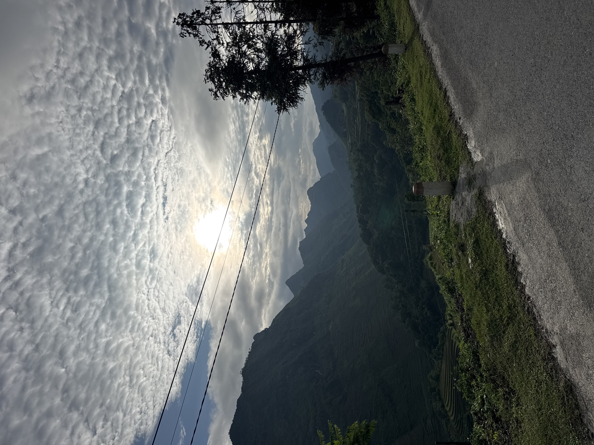 The image presents a rural landscape in Lũng Cú Commune, Vietnam. Mountains rise in the background, partially obscured by haze. A dense cloudscape covers the sky, with the sun positioned behind the clouds. Power lines are visible in the lower left. A road runs along the right edge of the frame, bordered by green vegetation. The scene appears serene, capturing the natural beauty of the mountainous region.
