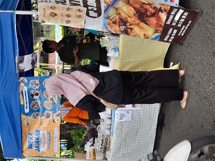 The image shows a food stall in Kecamatan Sidoarjo, Indonesia, located at coordinates -7.441 latitude and 112.705 longitude. The stall is set up under a blue canopy, with various food items displayed on the counter. One woman in a black shirt and pink hijab stands in front of the stall, facing the camera. She is looking directly at the viewer. Another woman wearing a bright orange shirt stands behind her. A man in a blue shirt is talking with someone, likely the customer, outside the stall. In the background, a third man in a blue shirt and black pants stands near the stall, looking at something to the right of the camera. The stall is set up on a paved street. It is adorned with a large banner advertising various food items, along with price lists for the food items. The banner mentions a price of "Rp 3,000," which is likely the price for the food being sold. There is a motorbike parked to the left of the stall, partially out of frame.