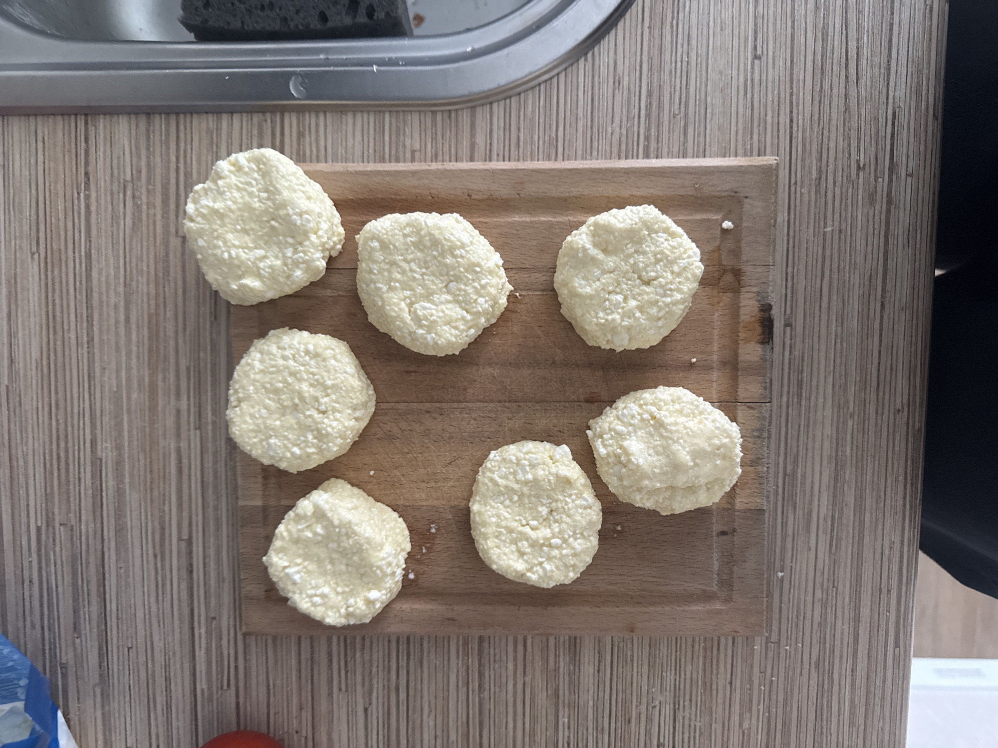 Seven round, pale yellow objects, possibly cheese patties, are arranged on a wooden cutting board. The cutting board is on a wood-grain laminate countertop. To the right of the countertop is a stainless steel sink. The lighting is overhead and casts soft shadows. The scene appears to be a kitchen. No information about the city of Perm, Russia is present in the image.