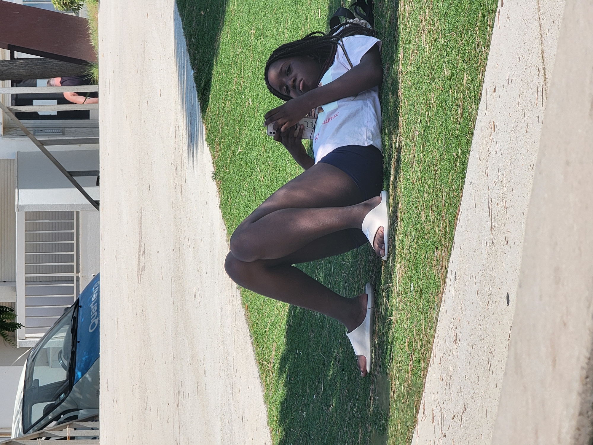 In the city of Quarteira, Portugal, a young Black female lies on her back on a grassy area, propped up on her elbows, and looking at a mobile phone. She is wearing a white t-shirt, dark shorts, and white slides. Her dark hair is styled in braids, some of which fall over her shoulder. She is positioned in front of a building with white walls and vertical metal railings. A small, dark-colored backpack is visible behind her. The ground in front of the grass is a light-colored concrete path, and a portion of a vehicle with a blue stripe and the word "Quarteira" on it is visible in the upper left corner. The lighting suggests it is daytime.