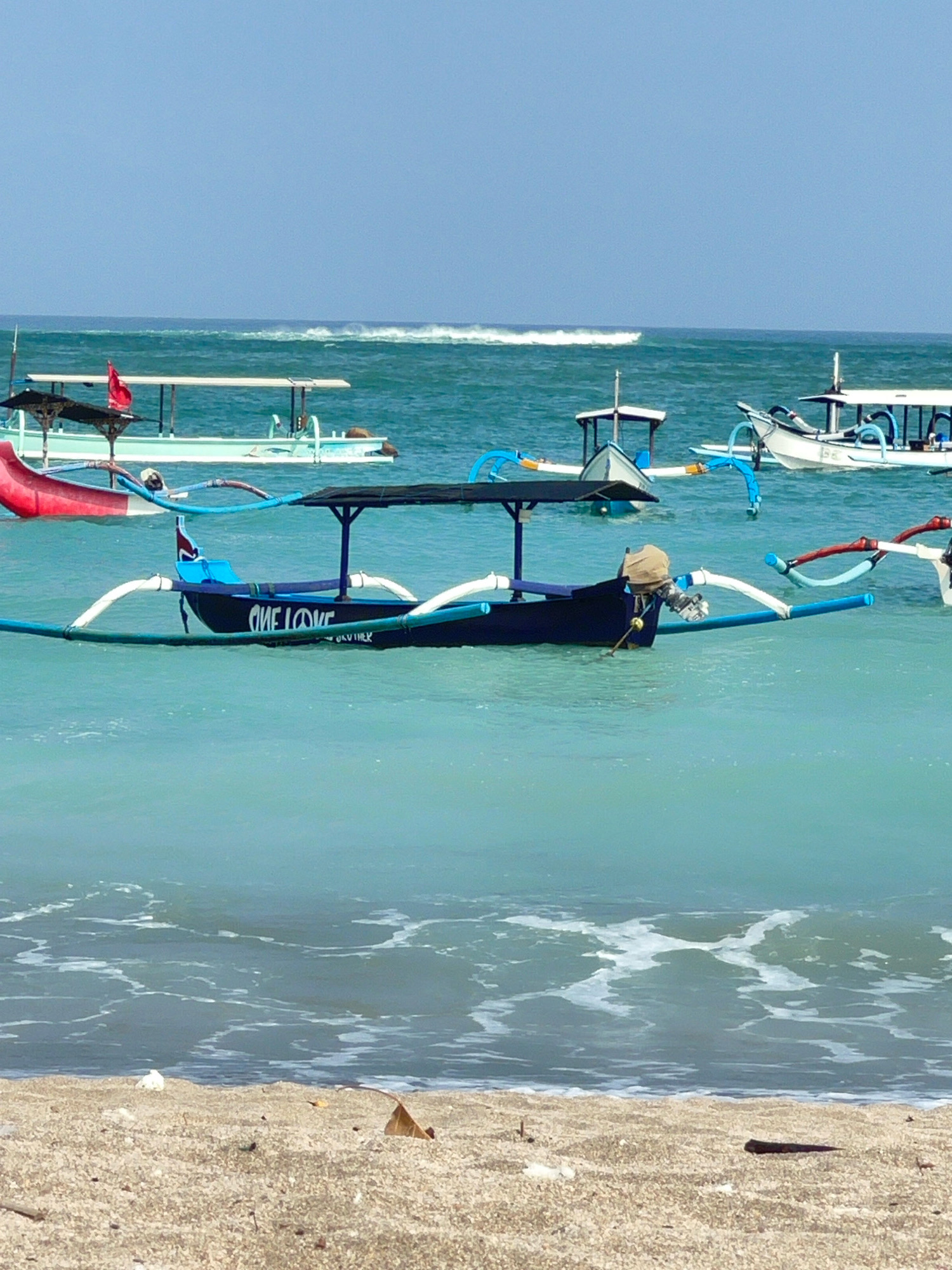 The image depicts a serene beach scene in Kuta, Indonesia, on a clear day under a pale blue sky. The foreground is occupied by a sandy shore with scattered debris, including a dried leaf and small white objects. Gentle waves with foamy white crests lap at the shore. In the mid-ground, several traditional Balinese jukung boats are anchored in the turquoise water. One prominent boat in the center of the frame is dark blue with "ONE LOVE" written in white on its side. It features a canopy supported by poles and outrigger floats. Other jukungs, varying in color and design, are visible further out, some with red flags or white canopies. In the background, the vast expanse of the ocean meets the horizon, with a faint white line suggesting distant waves. The overall impression is one of tranquility and coastal life in Kuta.