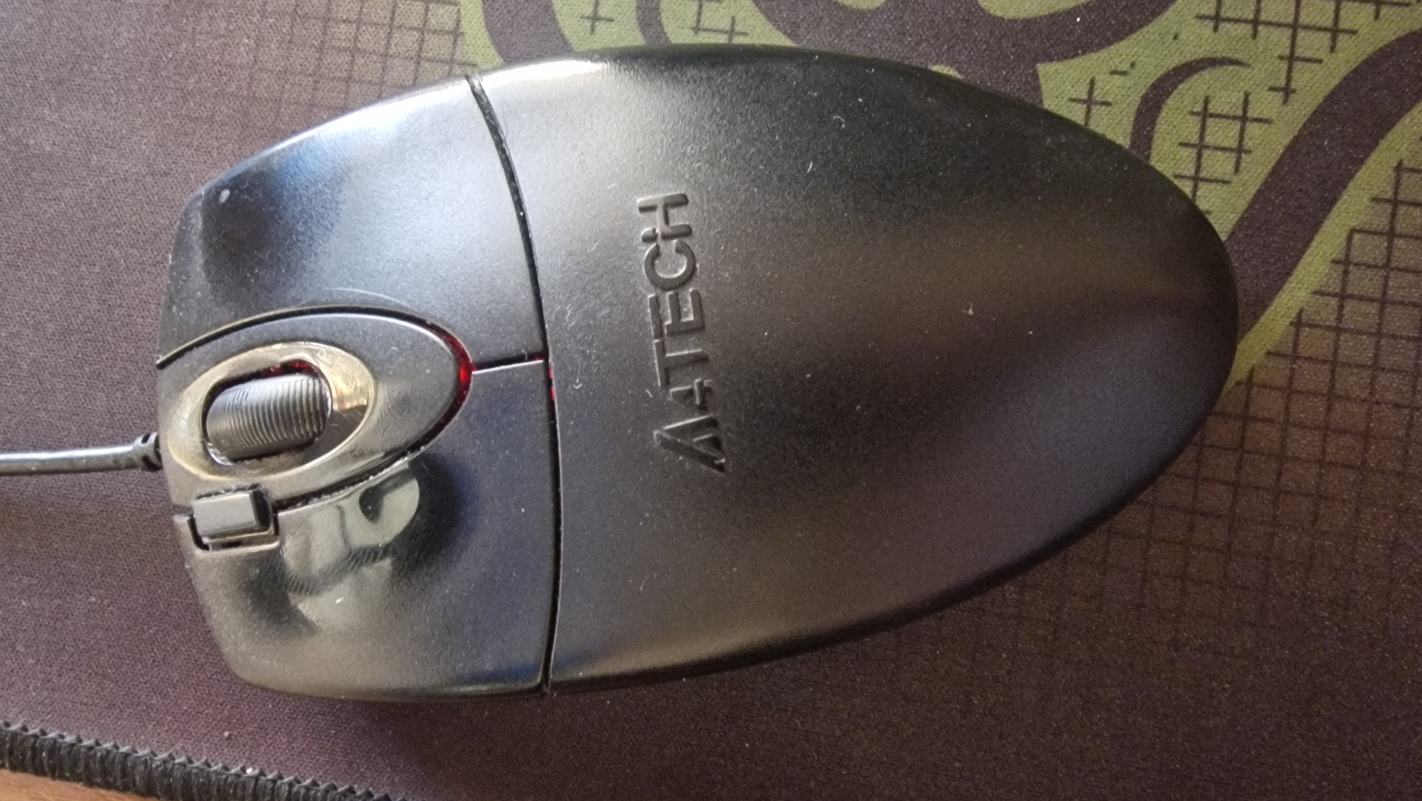 The image shows a black A4TECH computer mouse resting on a dark brown mousepad with a green and brown patterned design. The mouse is positioned in the center of the frame, angled slightly to the left. The top of the mouse features a scroll wheel and two buttons. The A4TECH logo is visible on the lower half of the mouse's body. The environment appears to be indoors, possibly a desk or table. There are no people present in the image. The lighting is adequate, with some reflections visible on the glossy surface of the mouse. There is no indication of the location being in Navoiy City, Uzbekistan.