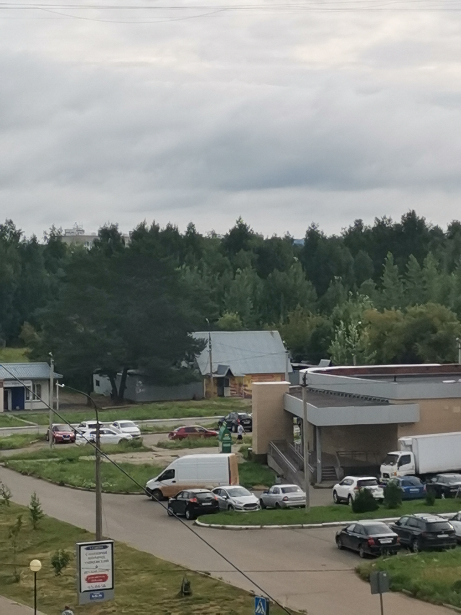 The image captures a cloudy day scene in the city of Chaikovsky, Russia, likely in a suburban or commercial area with residential buildings and a wooded background. In the foreground, a light-colored commercial building with a white awning and darker window frames is visible on the left. A white van is parked on a paved road. Further into the scene, a larger, light-colored building with a dark roof and a prominent entrance with stairs is situated. A white truck is parked next to this building. Numerous cars of various colors are parked in lot areas and on the road, including a red sedan, white sedans, a black sedan, and a dark SUV. The environment is characterized by green grass, trees, and asphalt roads. In the background, dense green trees form a backdrop with some taller residential buildings peeking through. A signpost with text and images is positioned on the left side of the image. The sky is overcast with diffuse light.
